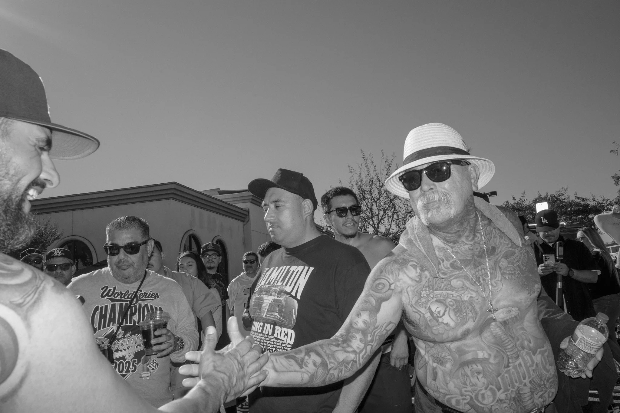 A group of men outdoors, some are shirtless with tattoos and wearing sunglasses and hats. One man is reaching out to shake hands with another. People in the background are observing, some holding drinks.