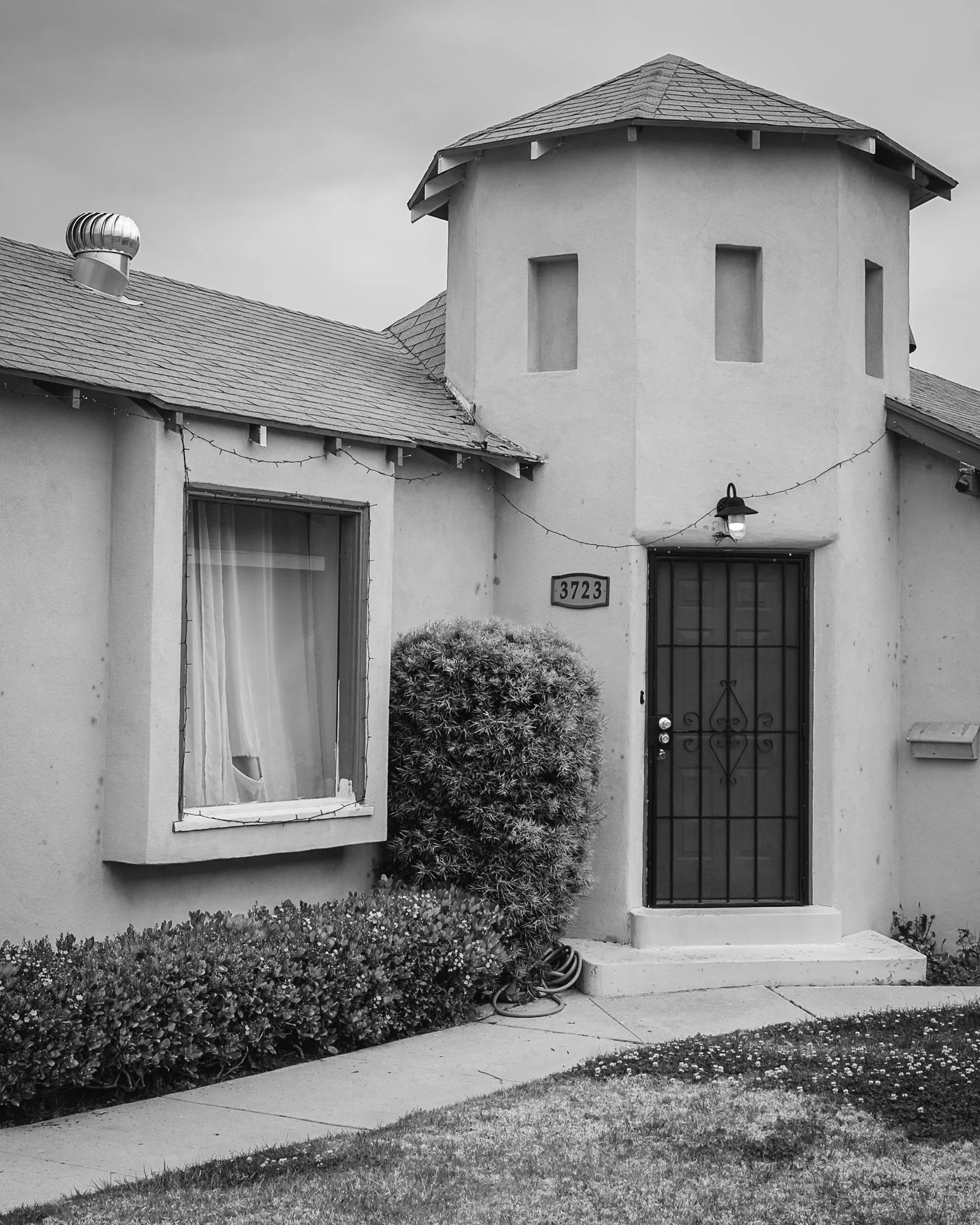 Black and white photo of a house with a turret, front door with a black security gate, window with curtains, bushes, and a sidewalk.