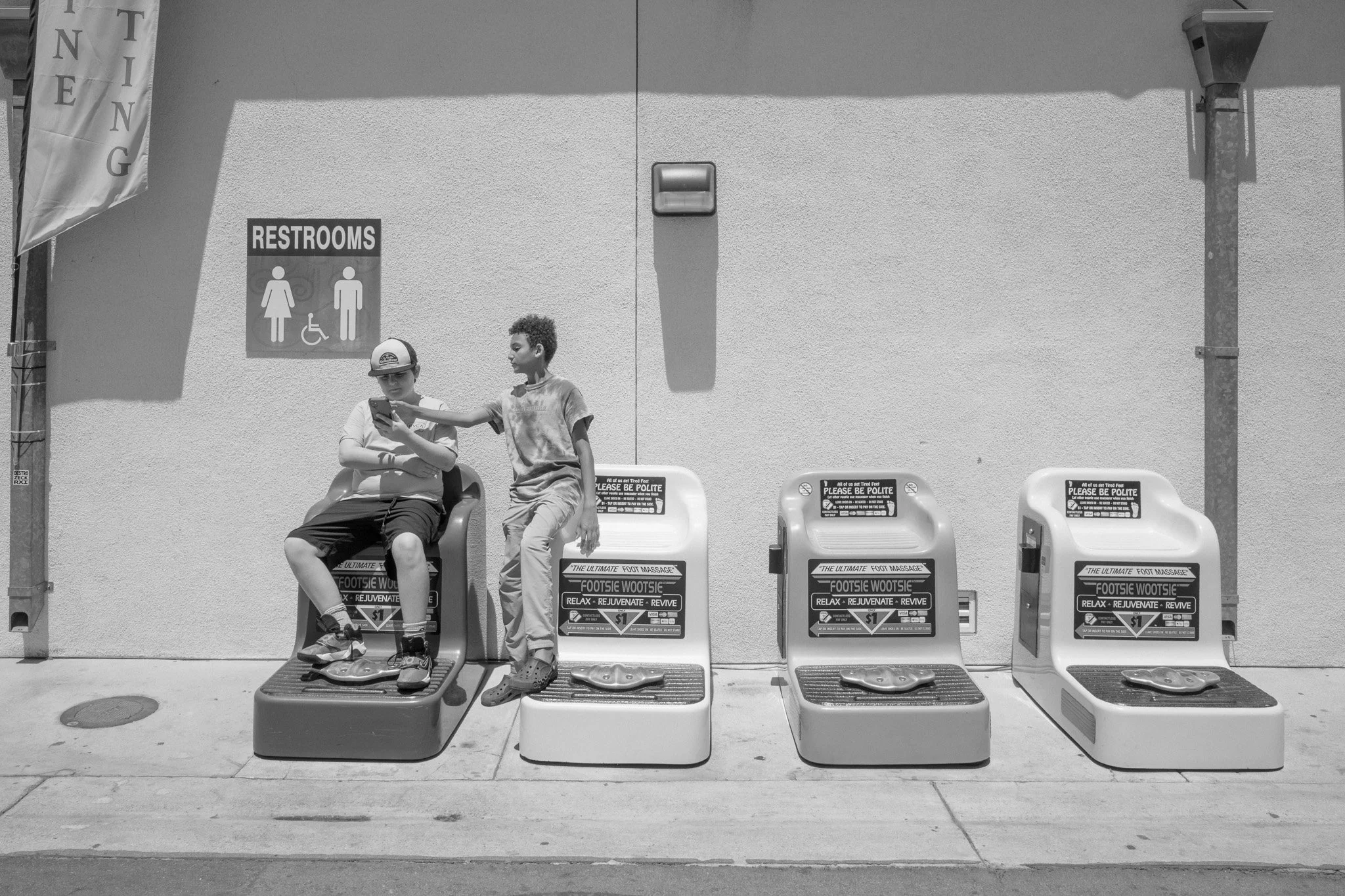 Two boys sitting and standing on foot massages machines outside a building with restrooms sign on the wall.