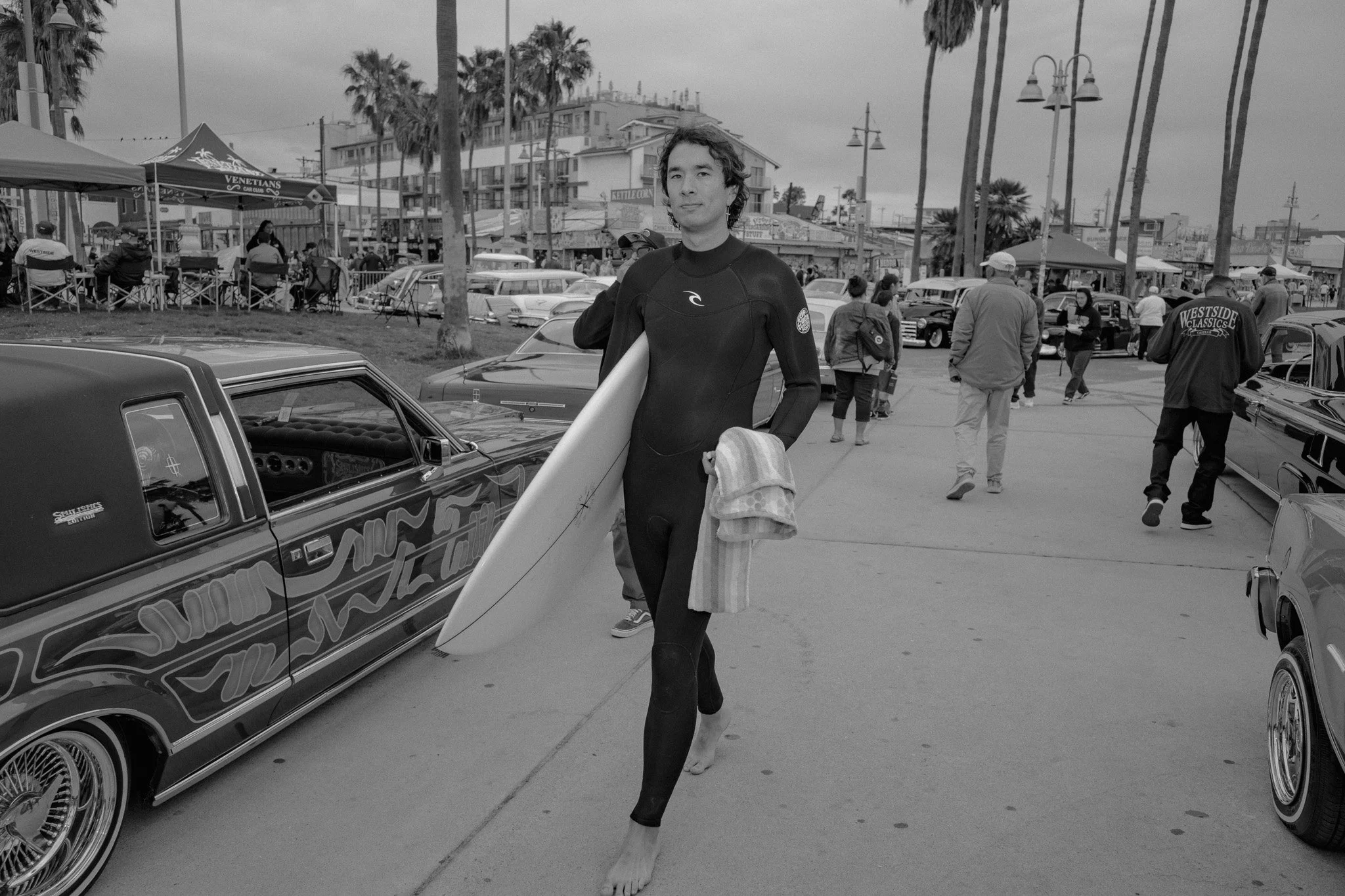 Person in a wetsuit walking on a sidewalk holding a surfboard and a towel, with classic lowrider cars and people in the background at a car show near the beach.