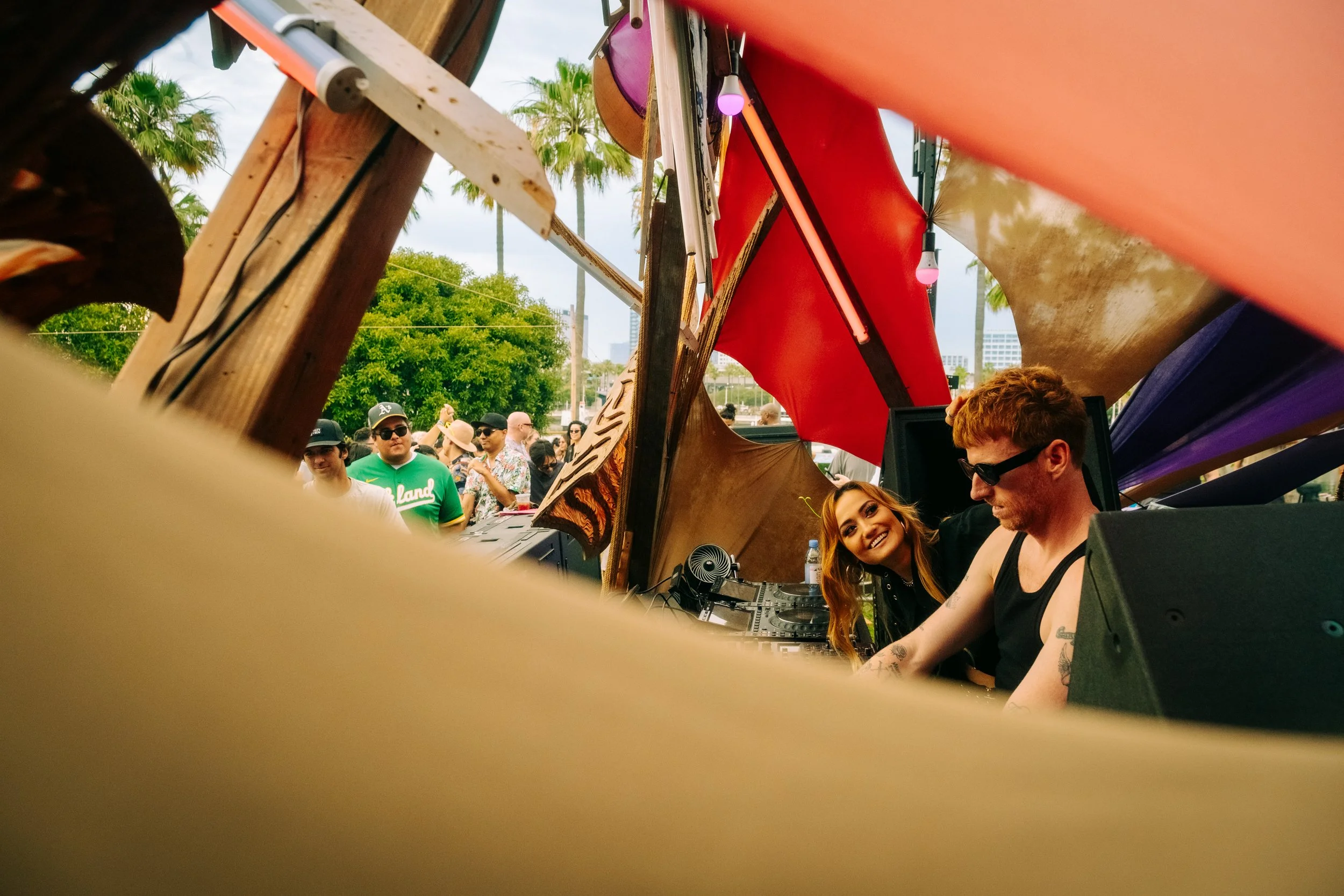 A DJ playing music at an outdoor event with a crowd and palm trees in the background. The photo is taken from a low angle, partially obscured by a blurred yellow object in the foreground.