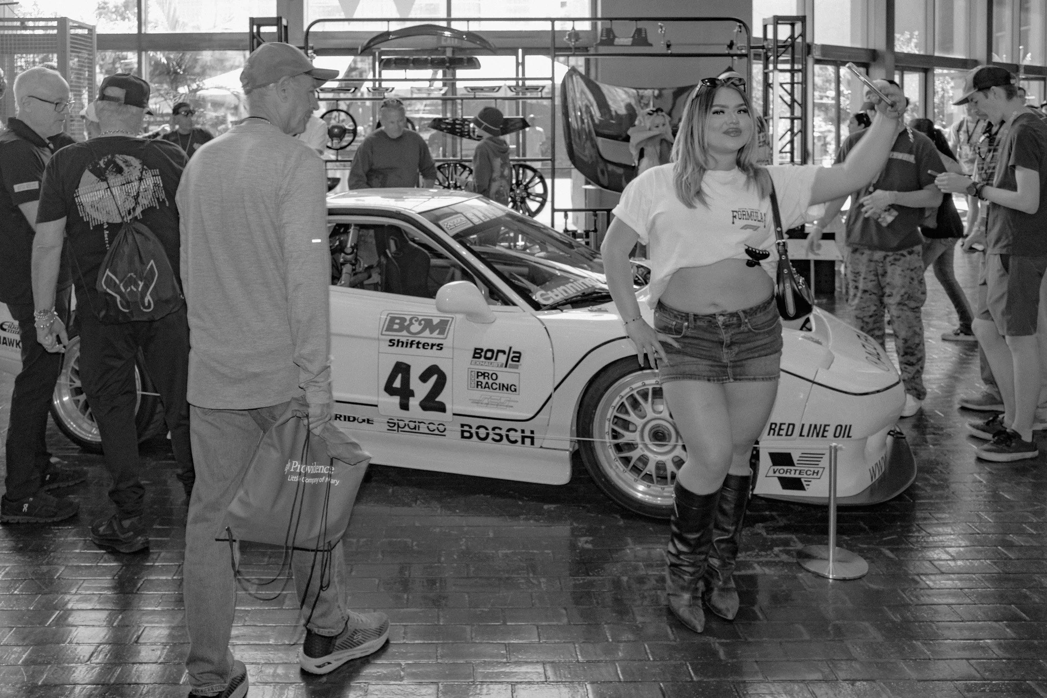 A woman in a white t-shirt, denim shorts, and knee-high boots is taking a selfie in front of a race car at an indoor car show. There are several people around, some looking at the car and others engaged in conversation.