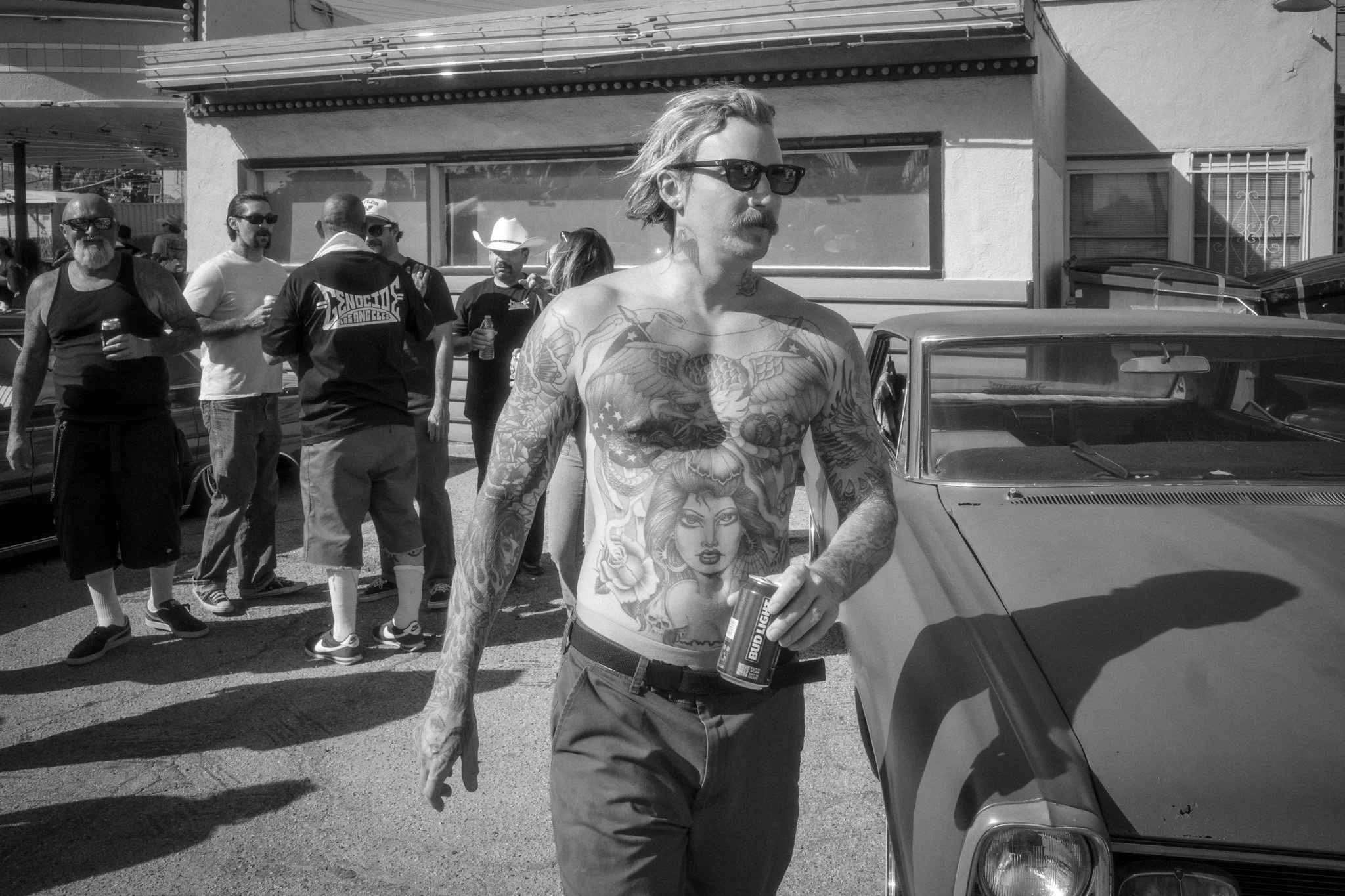 Black and white photo of a tattooed man with sunglasses holding a beer can, standing next to a vintage car, with a group of people in the background.