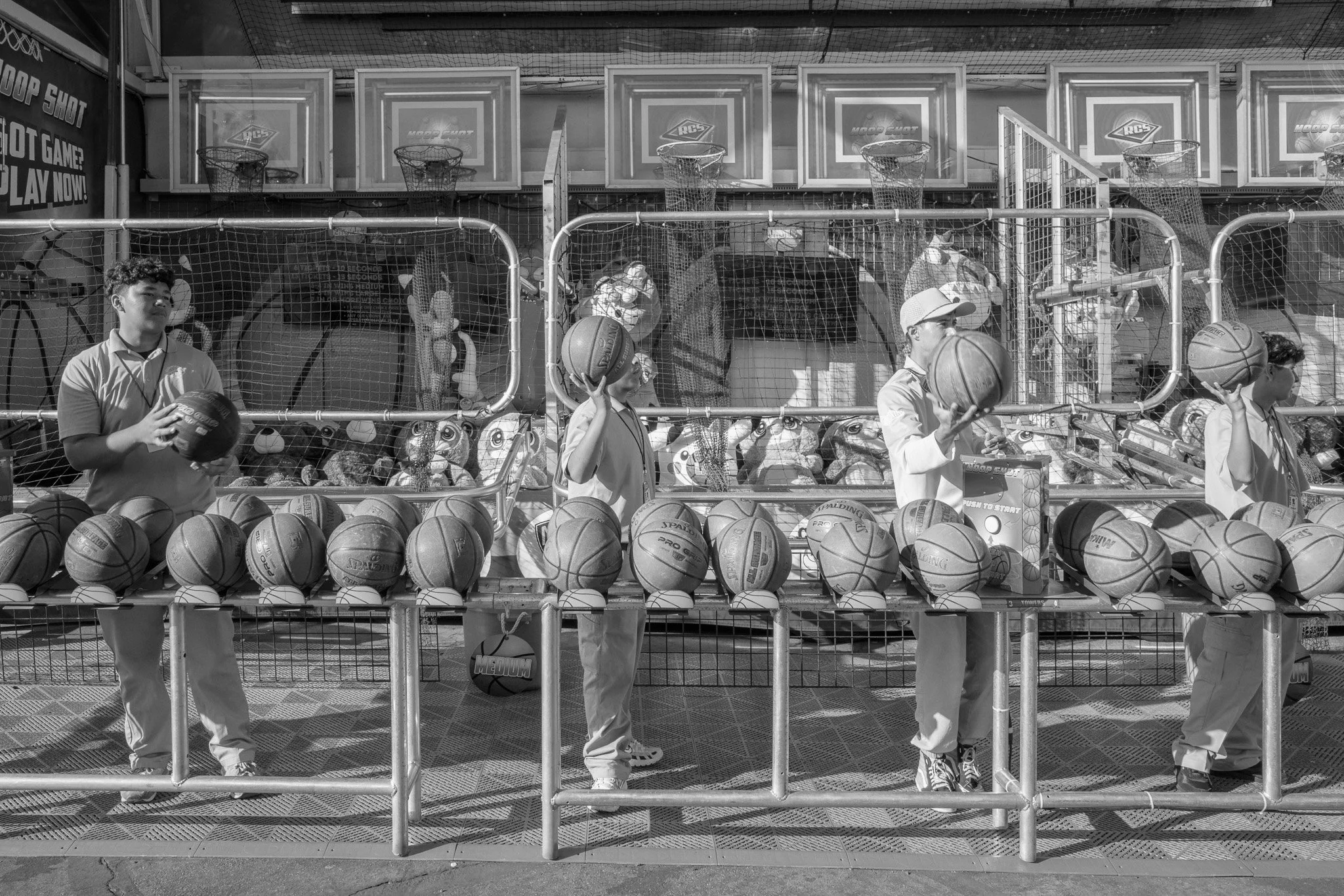 Four young boys at a basketball game shooting hoops at an arcade basketball game. They are holding basketballs, with many basketballs arranged on a table in front of them, and a fenced area with basketball hoops behind them.