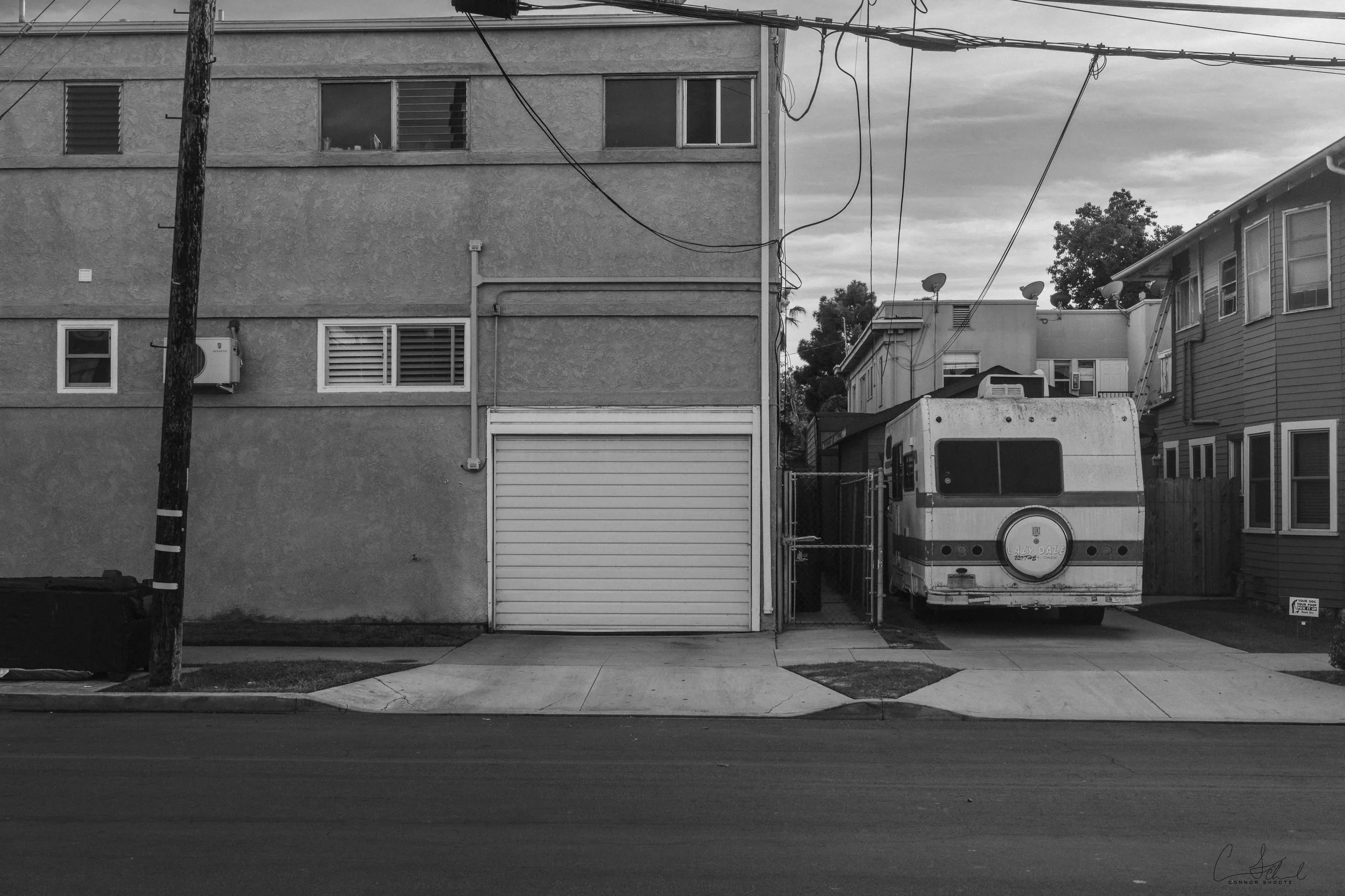 A black and white street scene showing a building with a garage door, a utility pole, a parked RV, and multiple power lines against a cloudy sky.