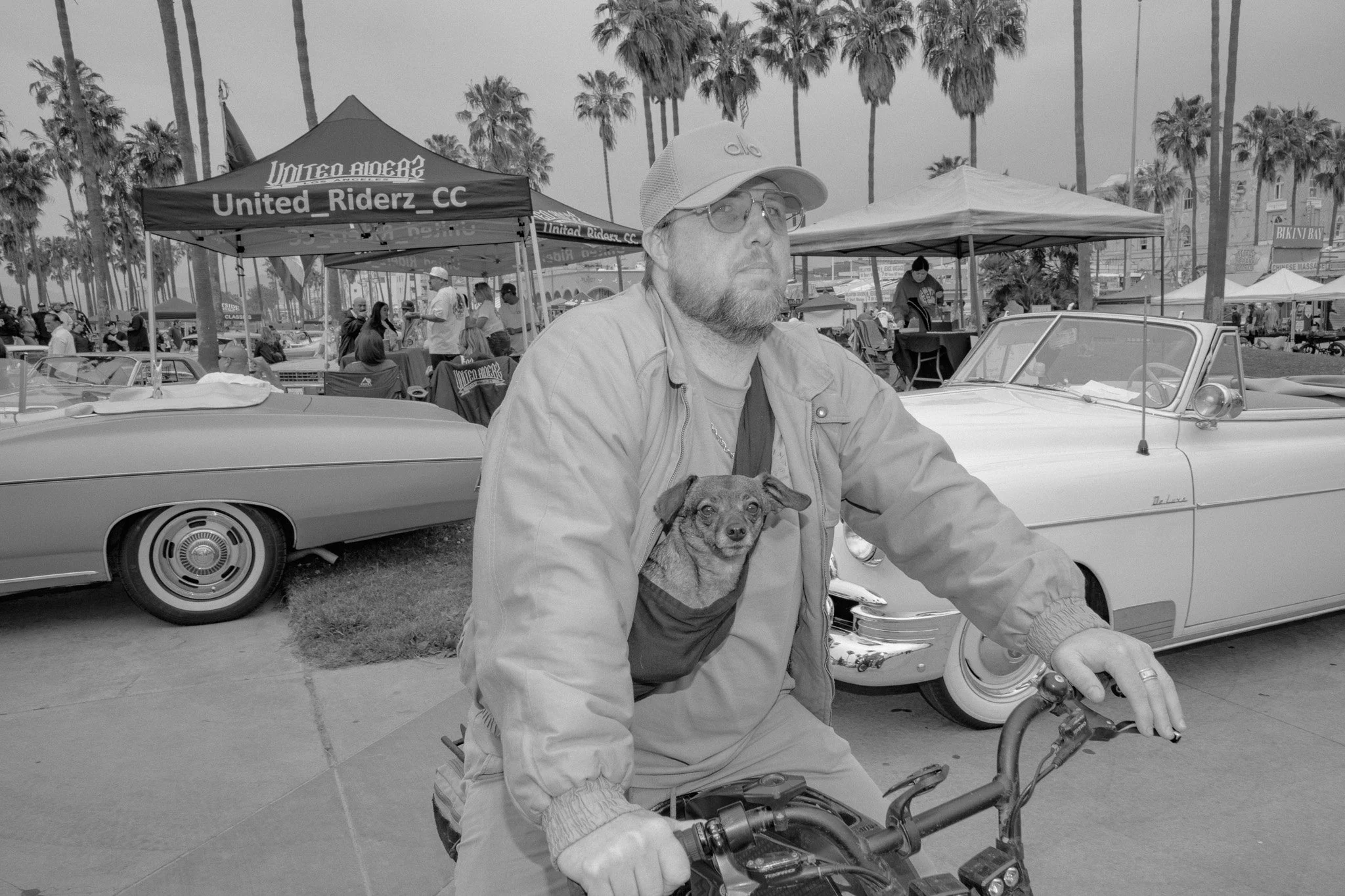 A man with a beard, glasses, baseball cap, and light jacket sitting on a vintage motorcycle with a small dog in a front chest pocket at an outdoor event with classic cars and palm trees.