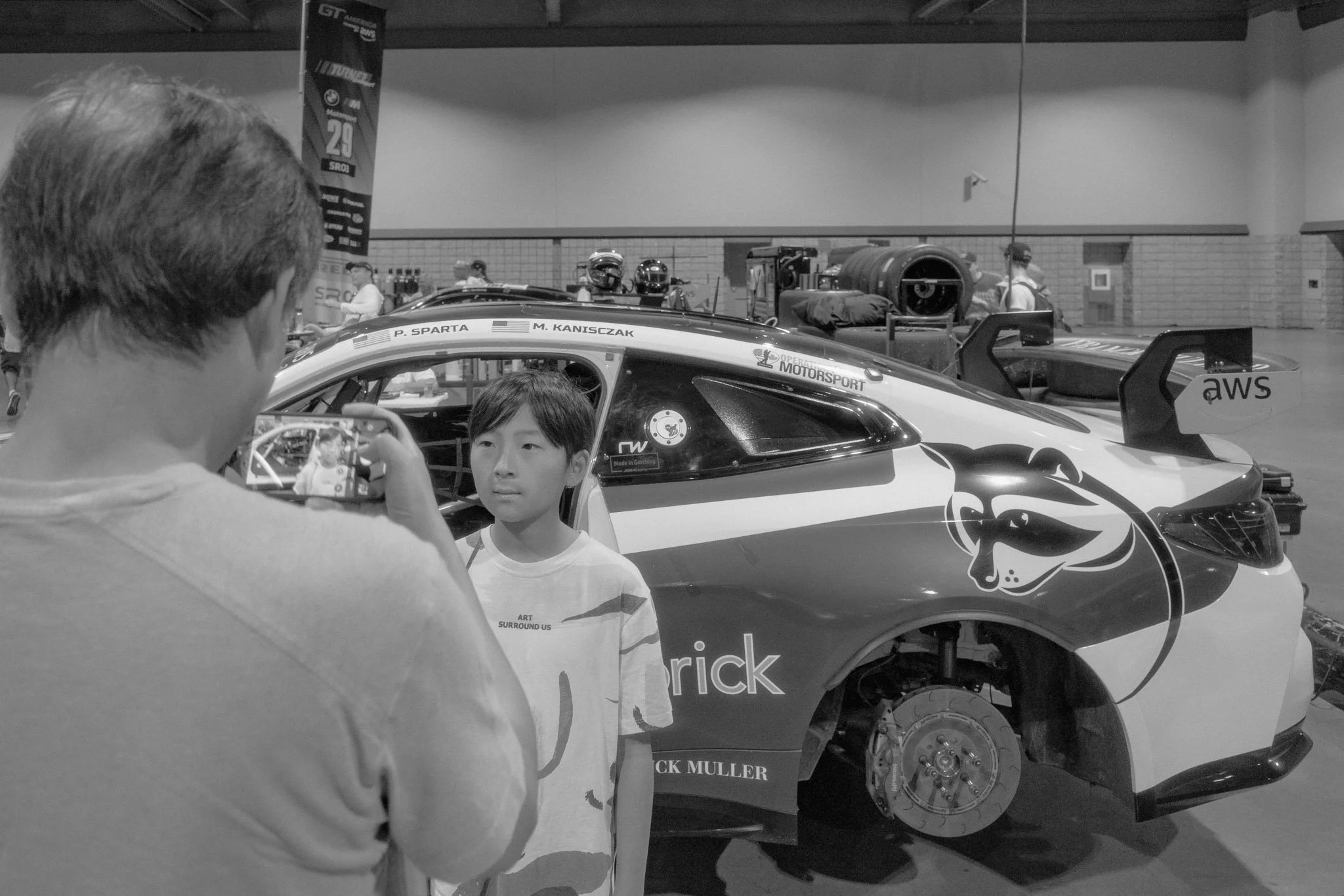 A dad takes a photo of his boy in front of a race car at the Acura Grand Prix in Long Beach. The boy is standing next to the car with various racing equipment visible in the background.