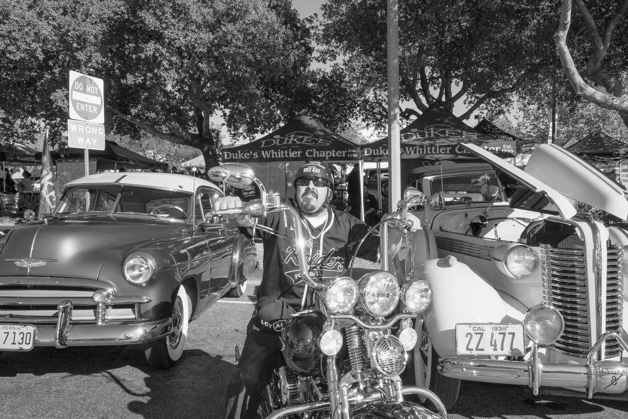 Man wearing sunglasses and helmet sitting on a motorcycle at a vintage car and motorcycle show. Classic cars and tents with signs in the background.
