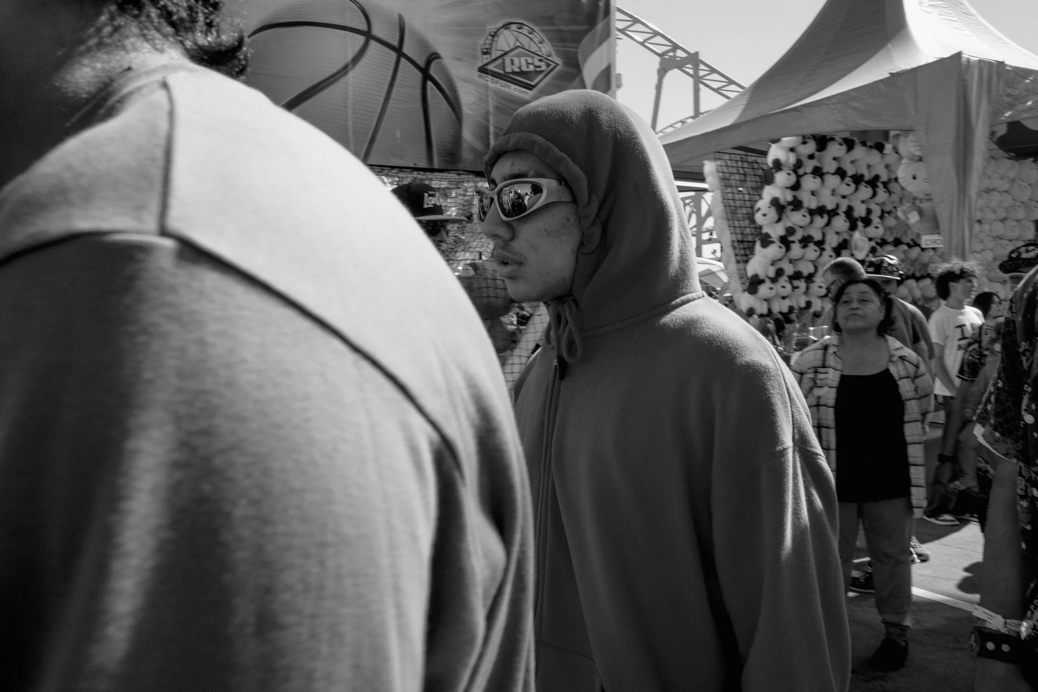 A young man wearing sunglasses and a hoodie stands in a crowded outdoor market or festival, with a booth selling stuffed panda toys and a sign featuring a basketball logo in the background.