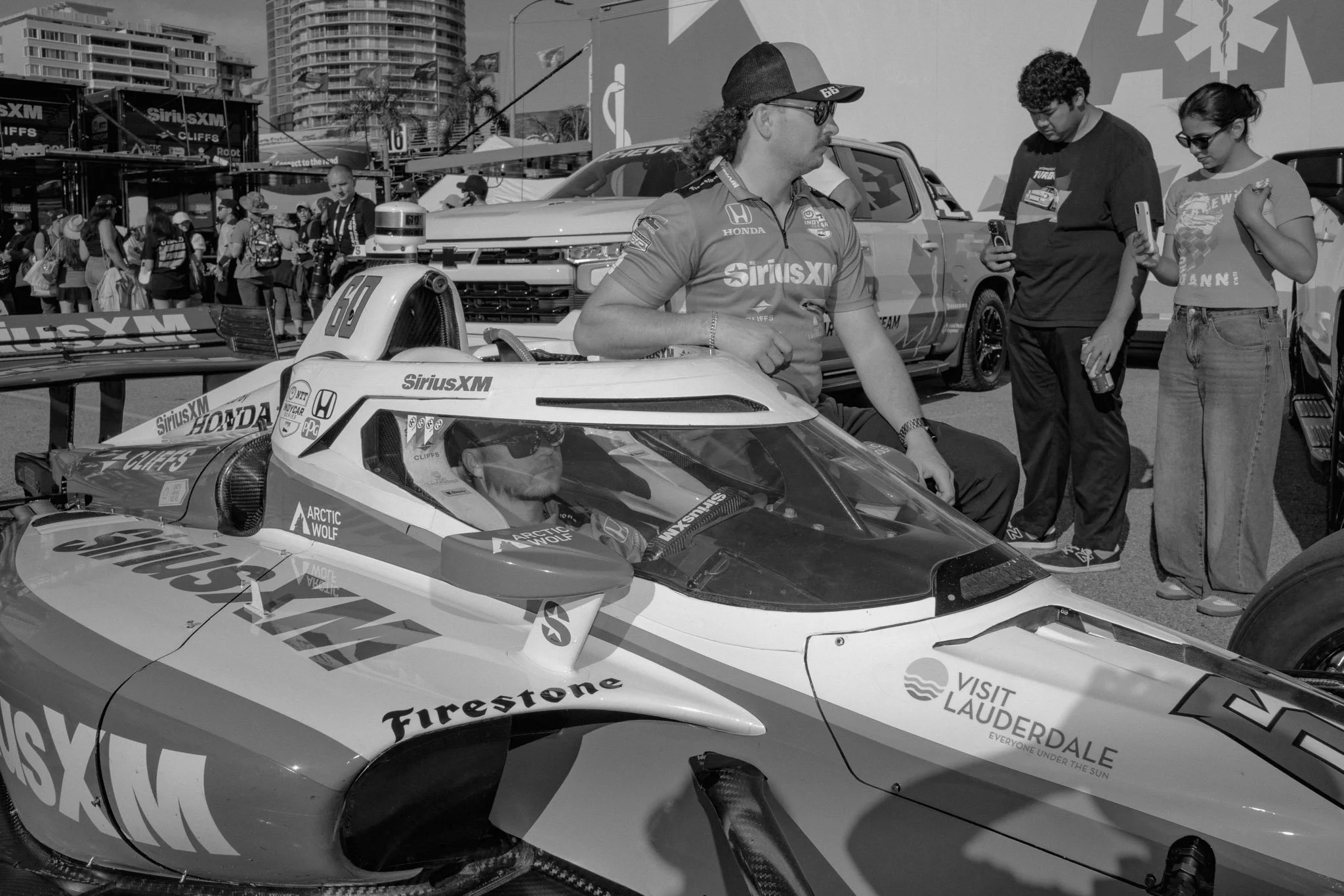 A person in racing gear sitting inside a race car with a group of people standing around outside at the Acura Grand Prix 2025 event, with buildings in the background.