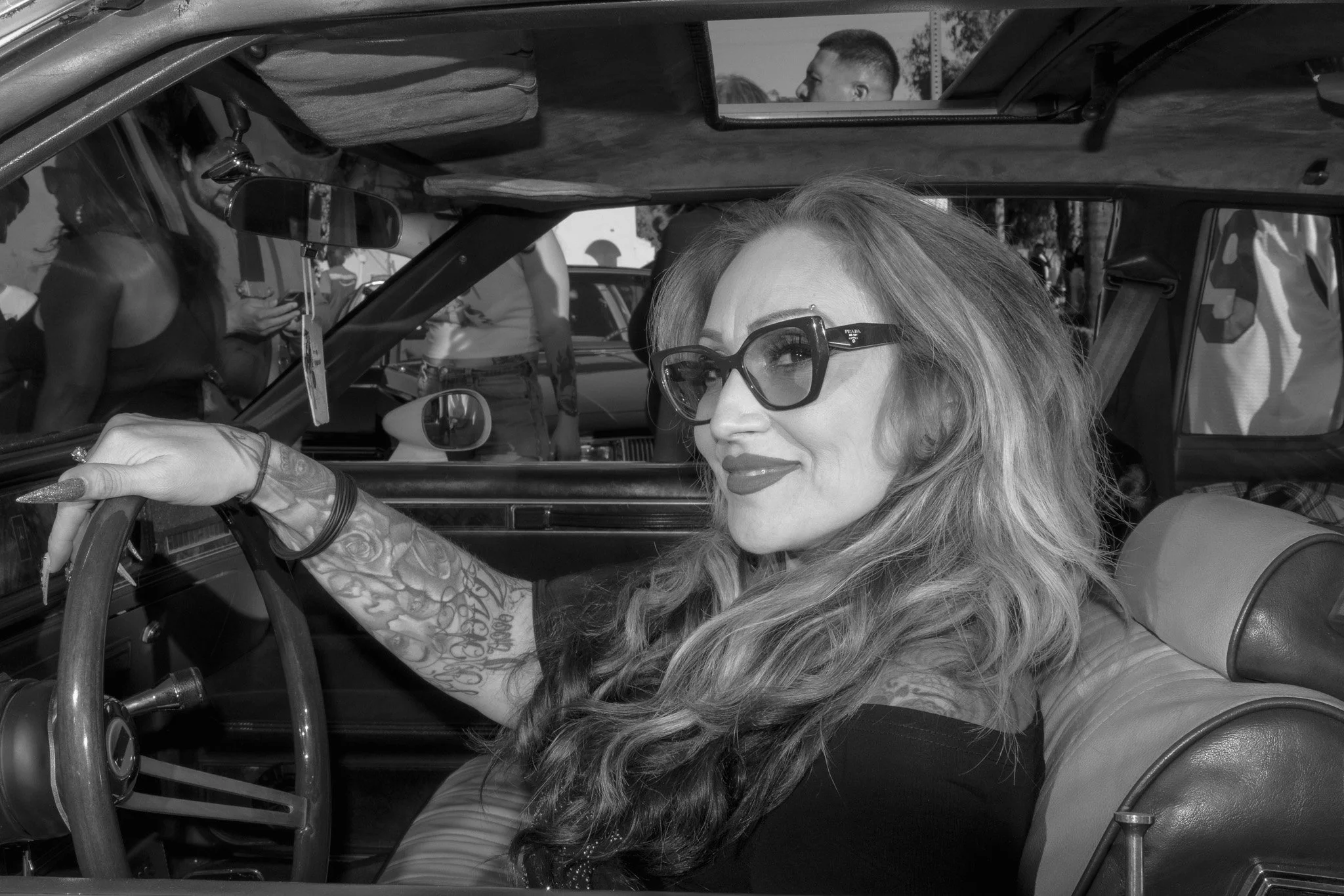 A woman with long wavy hair, wearing glasses, and a black top, sitting in the driver's seat of a vintage car. The woman is smiling and looking toward the camera.