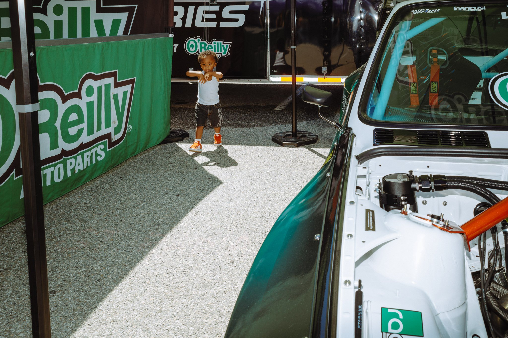 A young boy in a white tank top, black shorts, and orange sneakers standing next to a racing car at a motorsport event. The scene is set in a paddock area with banners and trailers in the background.
