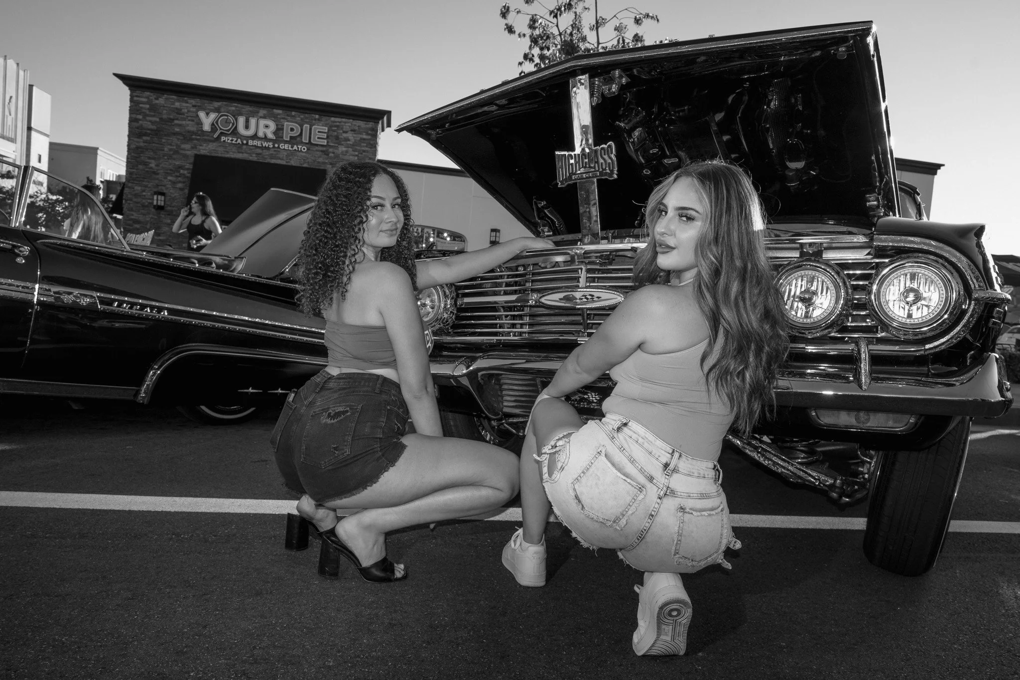 Two women with long curly hair and long wavy hair, both wearing crop tops and denim shorts, pose next to a lowrider with its hood open in a parking lot at dusk. A pizza restaurant sign is visible in the background.