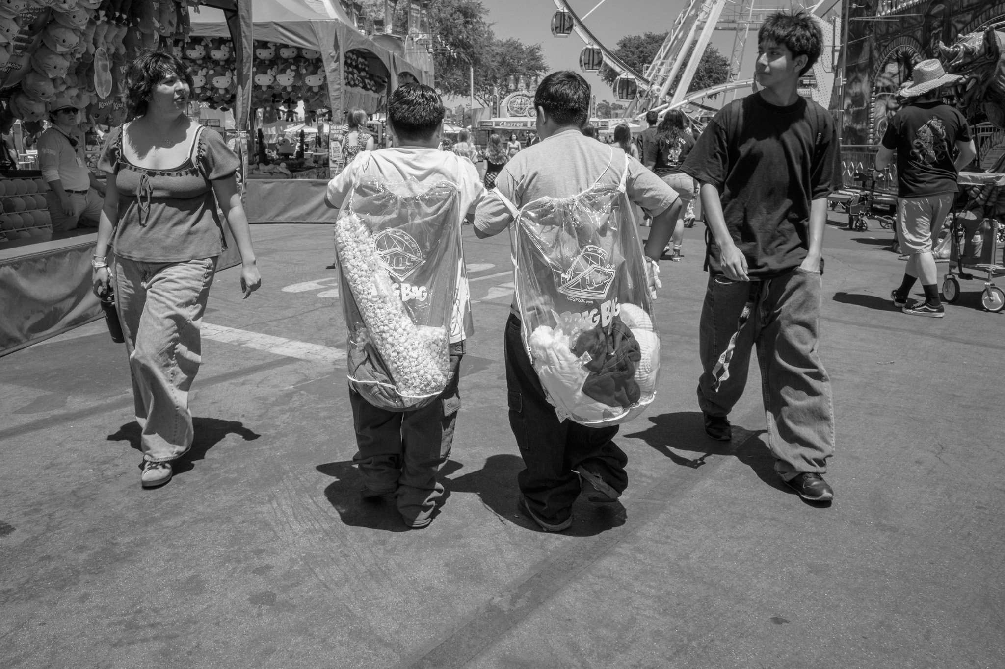 Four children walking in an amusement park, two with clear plastic bags filled with popcorn and other items, with rides and booths in the background.
