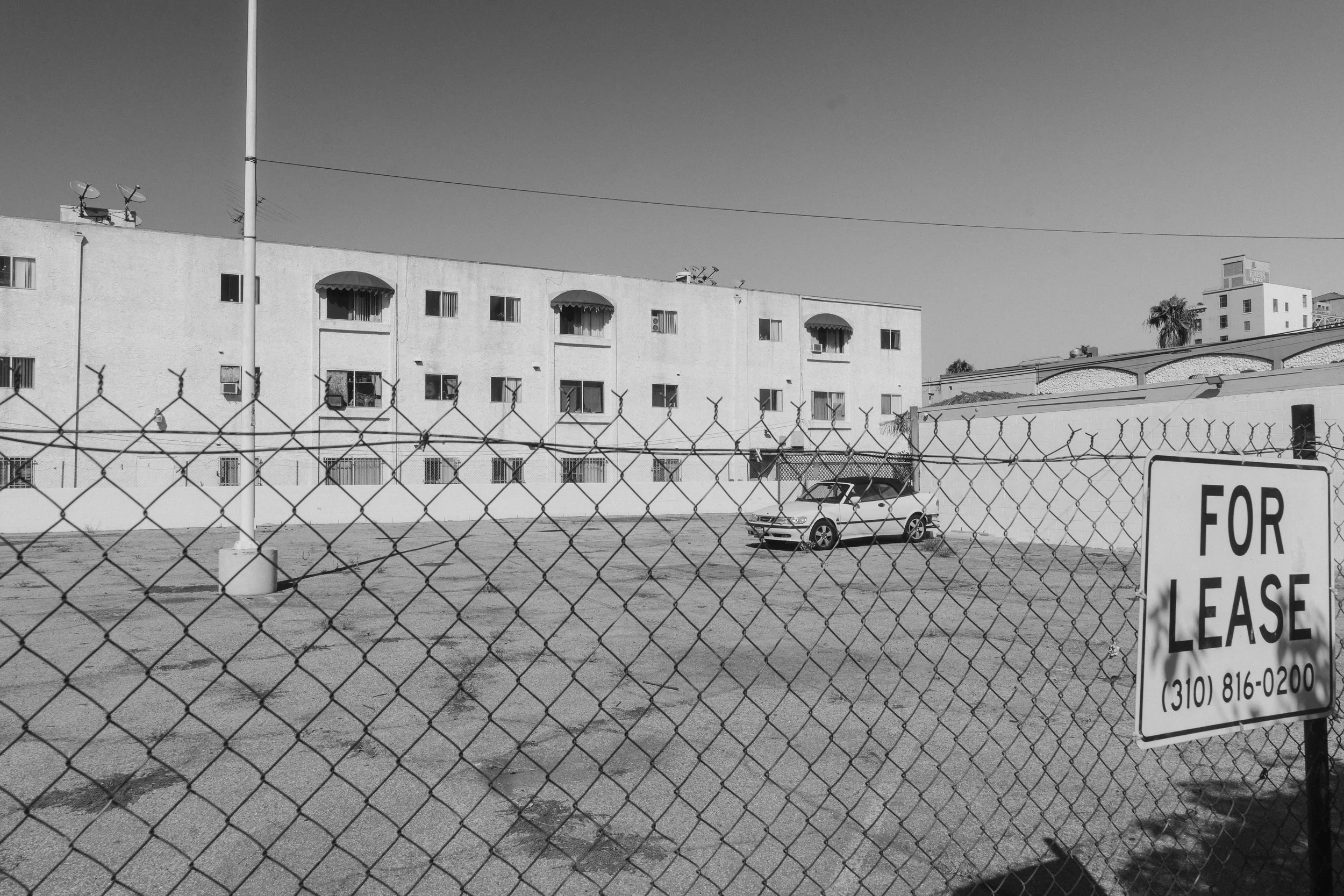 Empty parking lot behind a chain-link fence with a 'For Lease' sign, a white car, and an apartment building in the background.