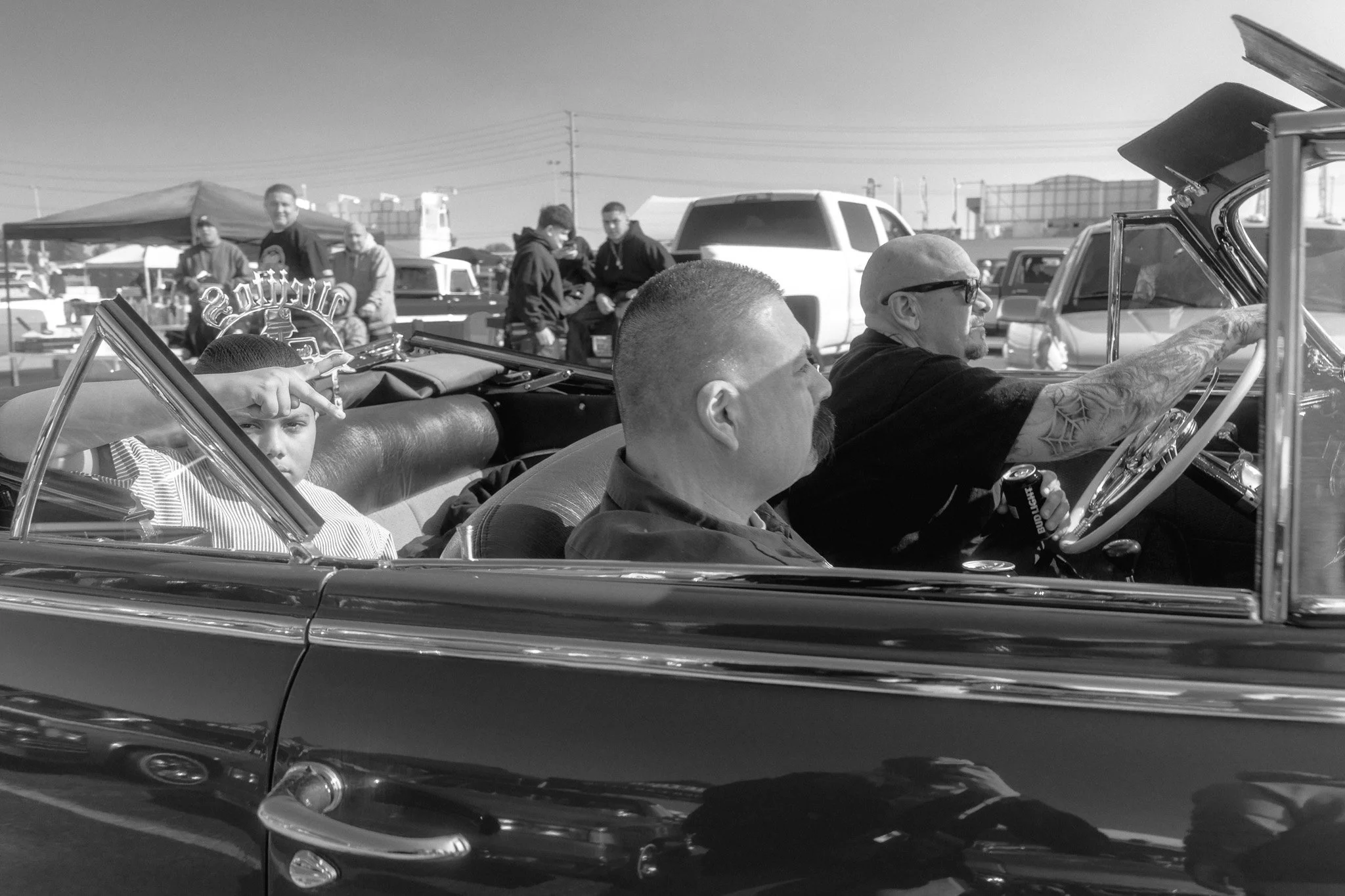A black and white photo of a vintage convertible car with three passengers; a young boy in the back seat making a peace sign, and two adult men in the front, one driving and the other seated beside him, at a car show or gathering.