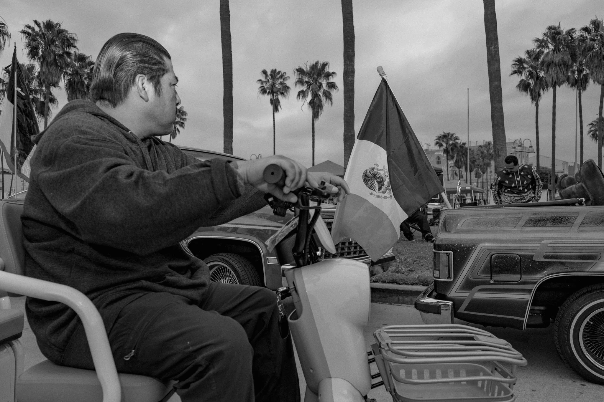 A man riding a mobility scooter with a Mexican flag attached in a parking lot. Tall palm trees and a person in patterned clothing are visible in the background.