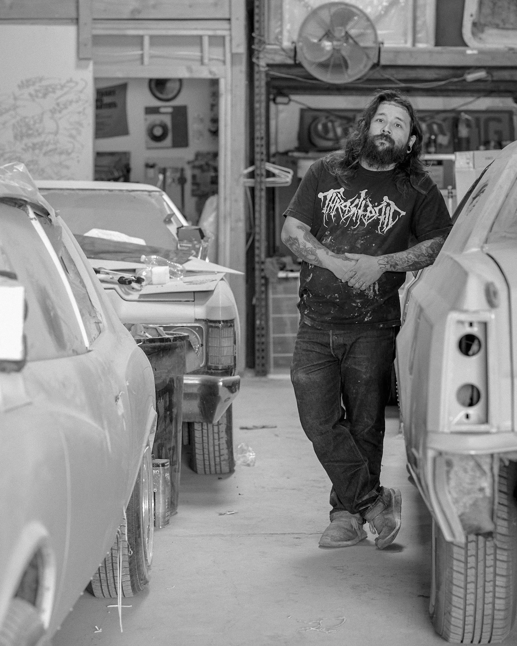A man with long hair and a beard standing between two vintage cars in a garage or workshop, with shelves and tools in the background.