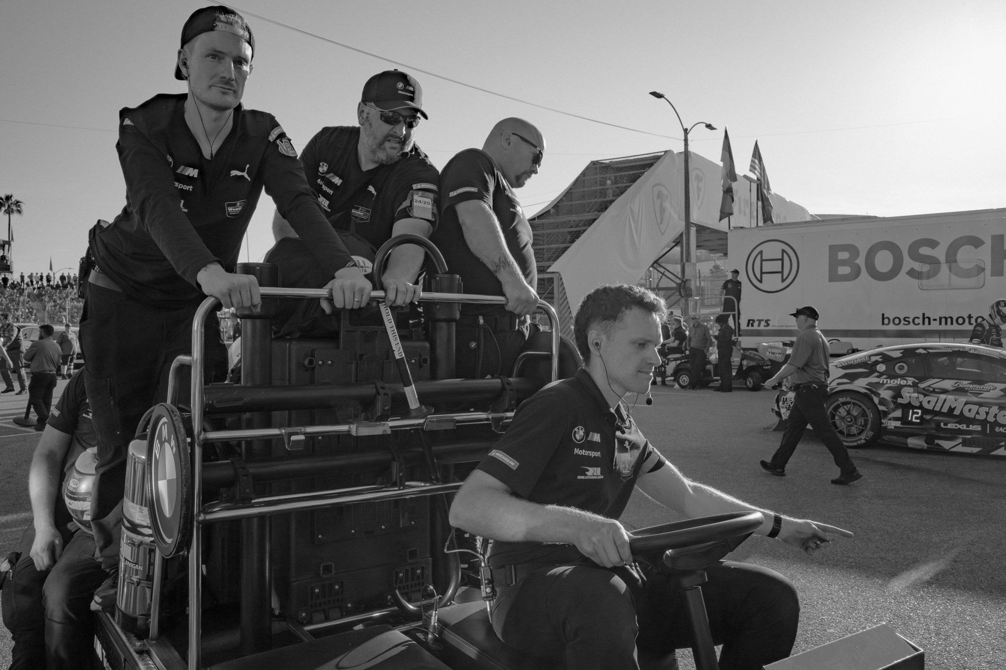 A team of IndyCar mechanics and a driver in a racing paddock, with cars and equipment in the background, some people walking around, and a large Bosch banner.