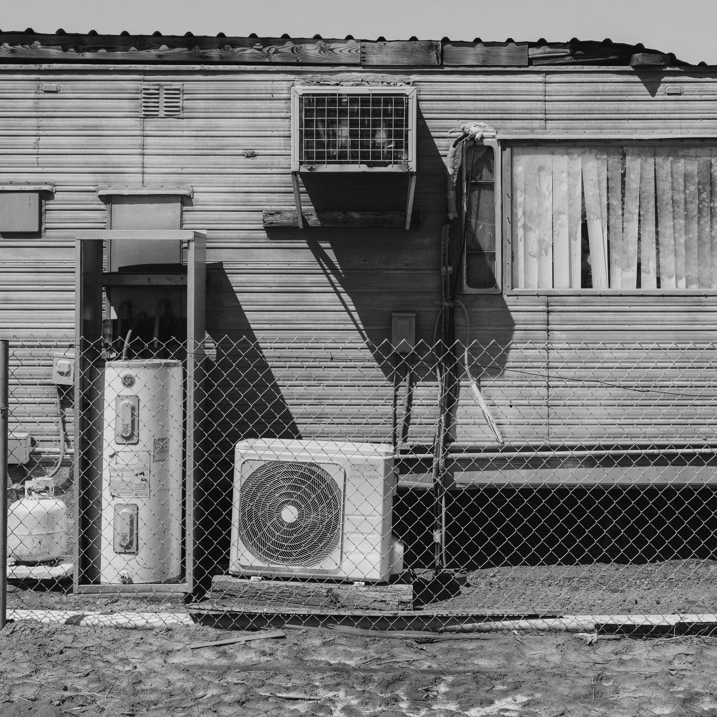 A black and white photo of an old mobile home with a window covered by curtains, an air conditioning unit, and a chain-link fence in front.