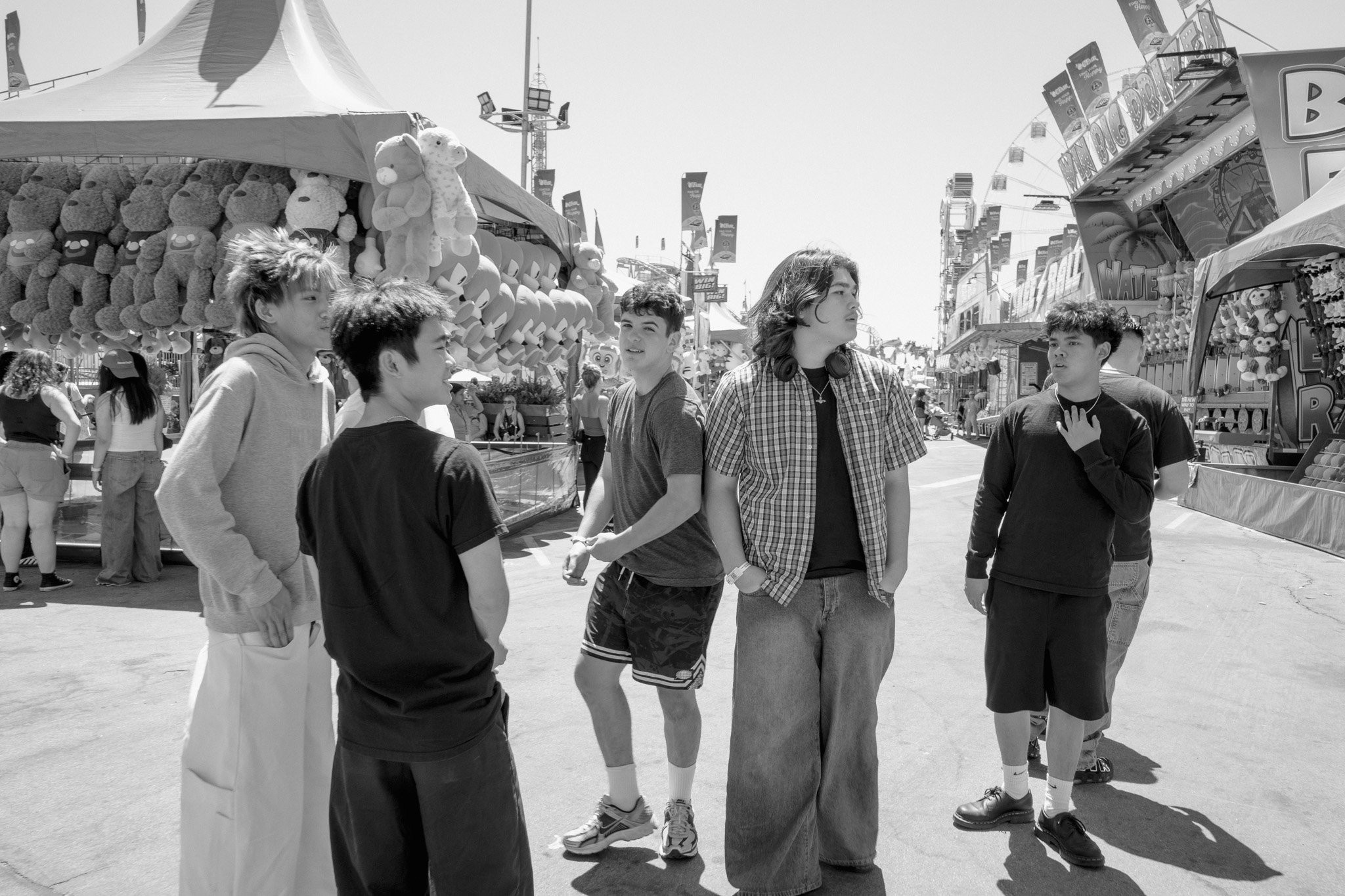 A group of five teenagers standing and talking at an amusement park or carnival, with game booths and rides in the background.