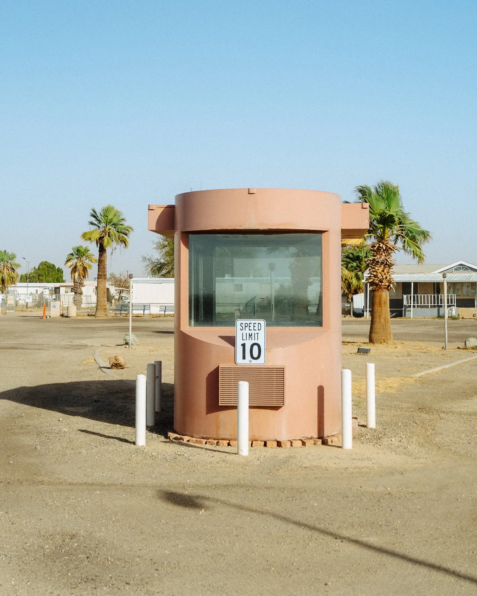 Small pink booth with a large window, white speed limit sign reading 10, surrounded by white bollards, desert landscape with palm trees and mobile homes in the background.