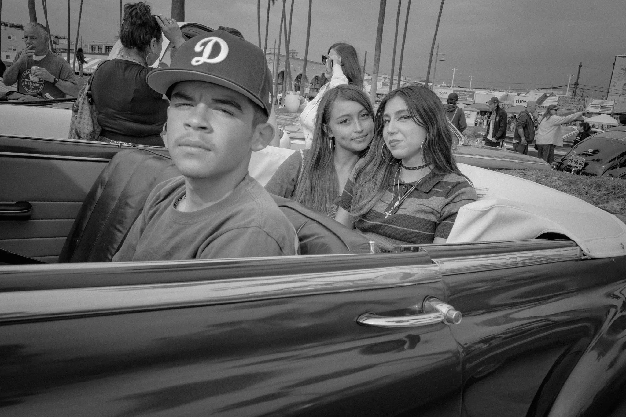 Three young women sitting in the backseat of a convertible car, with one young man in the front seat, at an outdoor event or fair, with people and tents in the background.