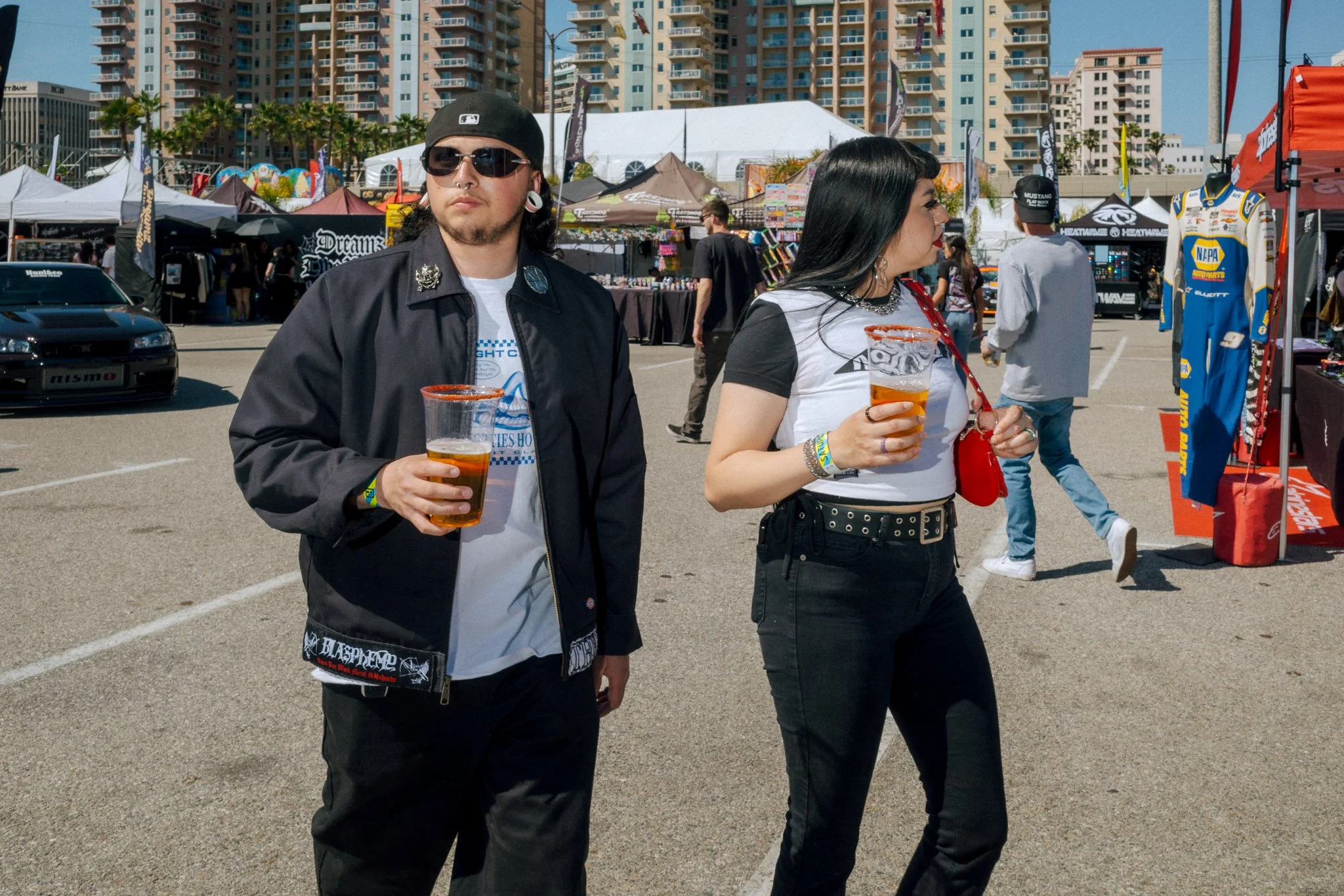 Two people walking at an outdoor event or festival, each holding a cup of beer, with tents, booths, and tall buildings in the background.