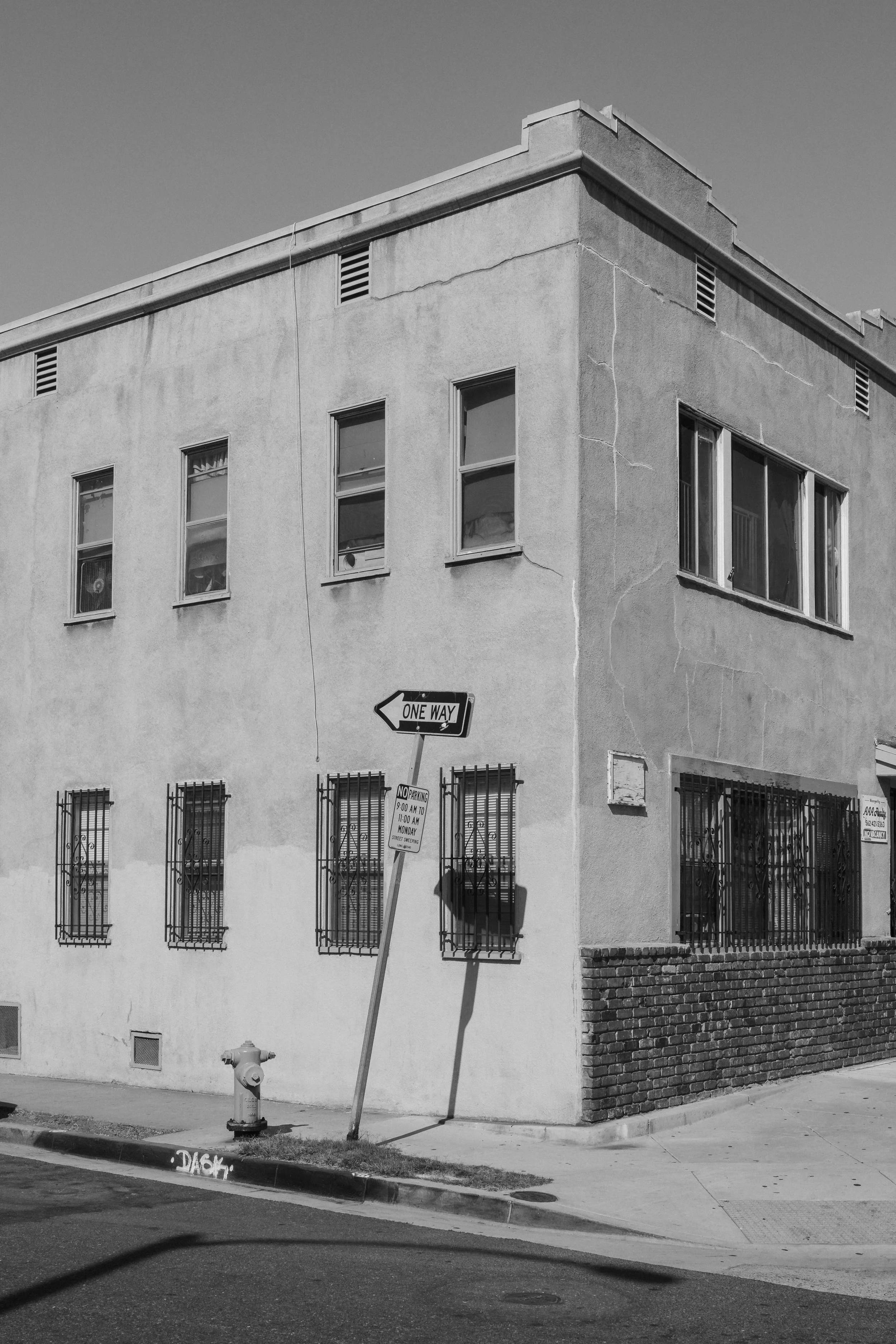 A black and white photo of a three-story building with barred windows on the lower level, a cracked exterior wall, and an end corner visible. There is a one-way street sign, a no parking sign, and a fire hydrant on the sidewalk.