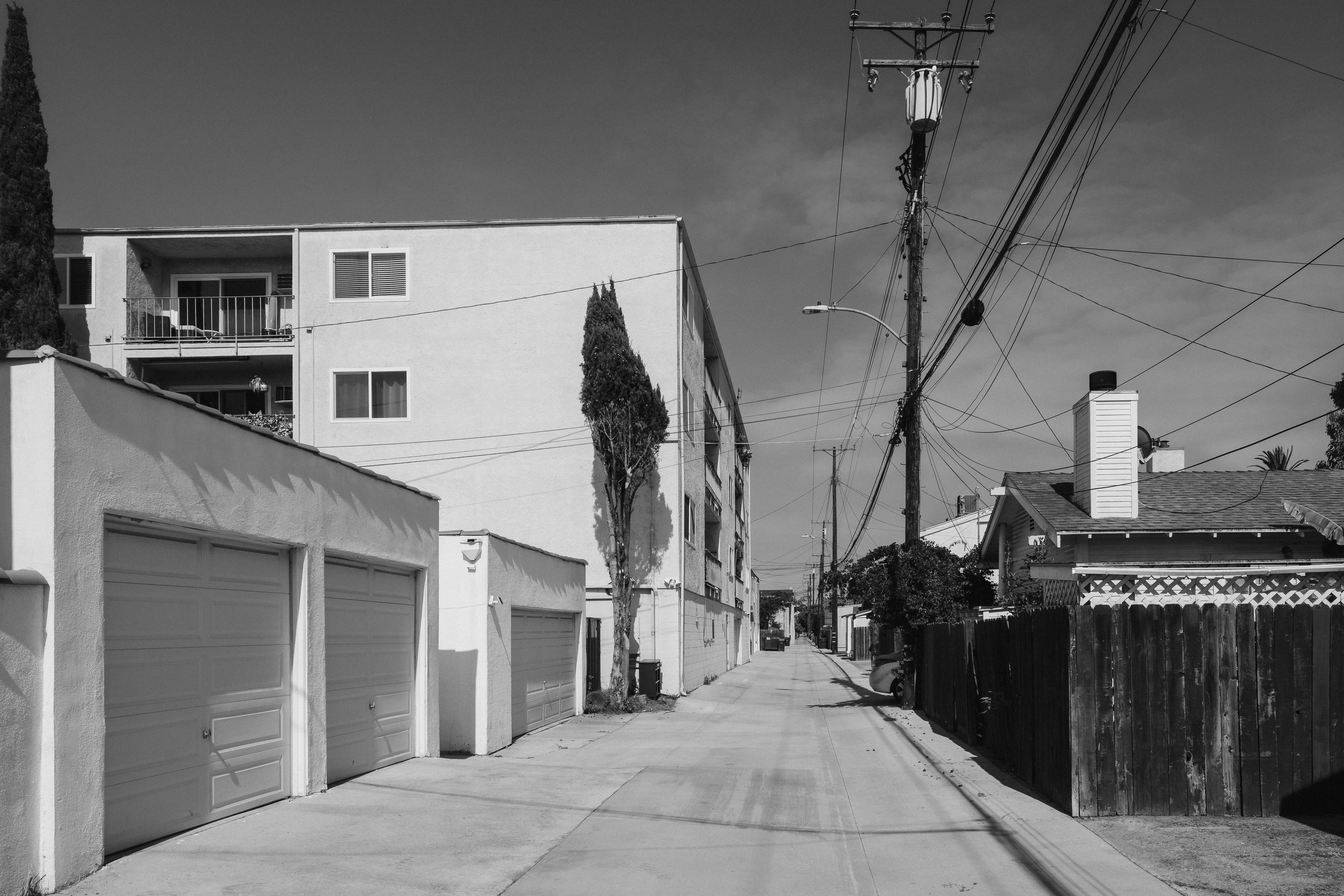 A black and white photo of a quiet residential street with garages on the left and houses on the right. Power lines and utility poles line the street, with a tree visible in the foreground and rooftops in the background.