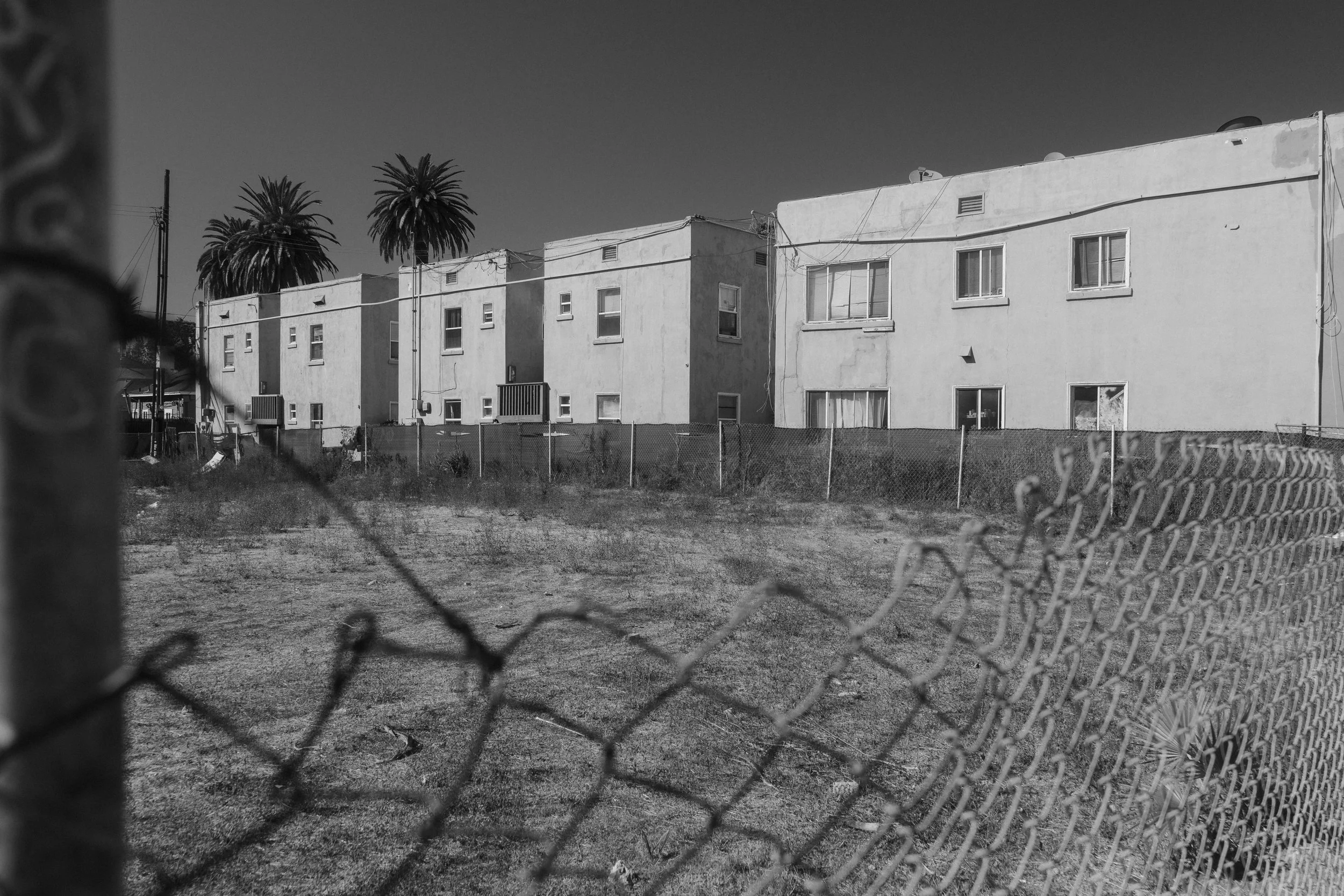 Black and white photo of an apartment complex behind a chain-link fence, with palm trees in the background, under clear sky.