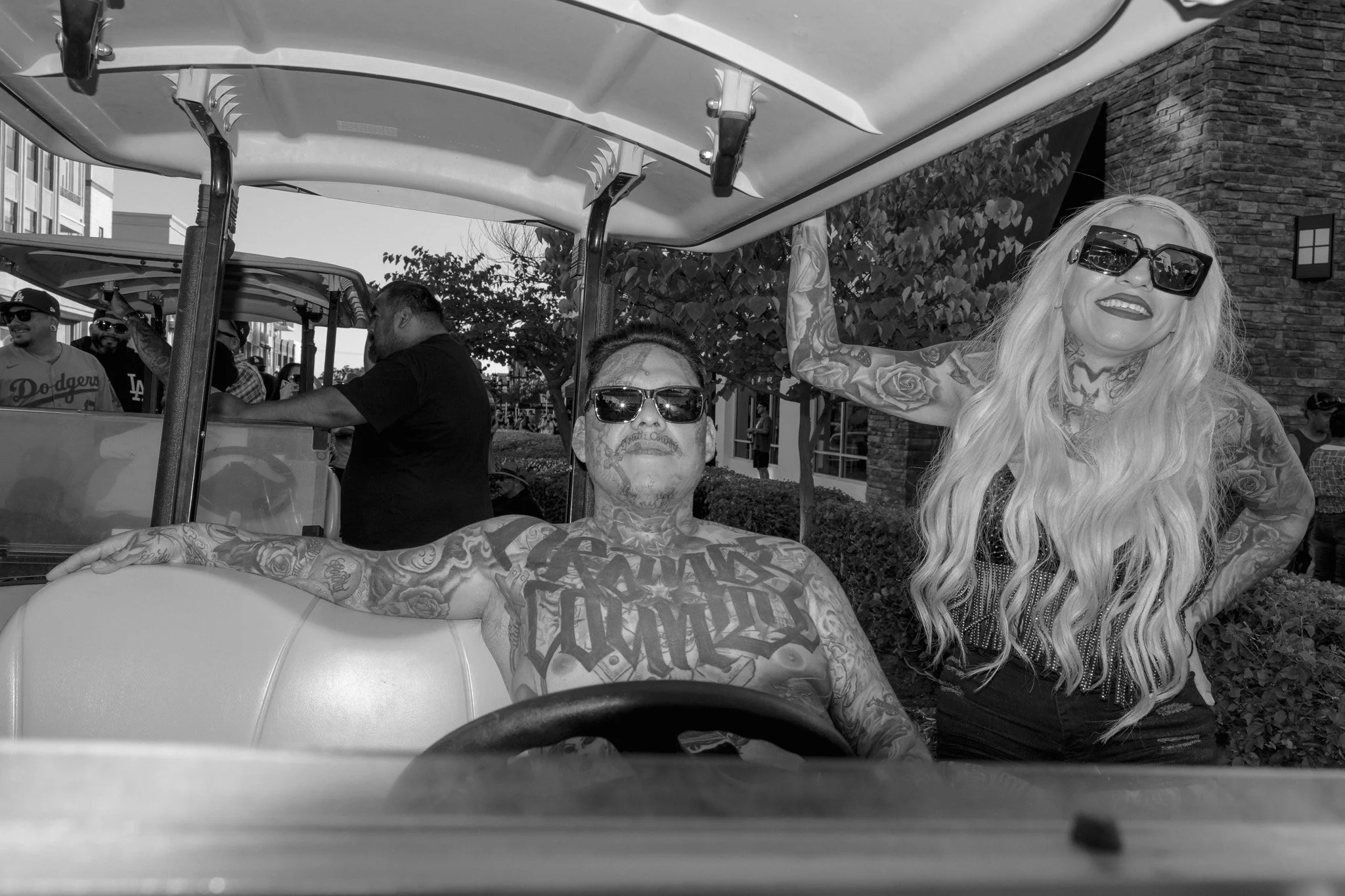 A man and a woman with tattoos wearing sunglasses riding in a golf cart during a parade. The man is sitting in the cart, while the woman stands beside him with her arm raised, posing for the camera. There are spectators and other golf carts in the ba