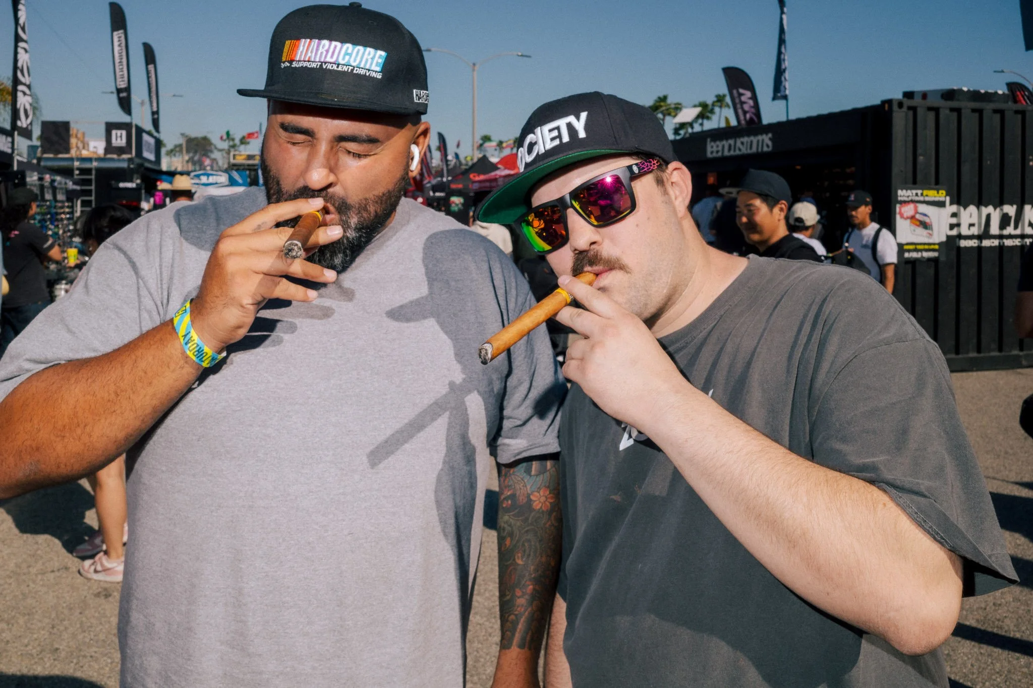 A portrait photograph of two young adult men with cigars at a formula drift car racing event. The men are spectators wearing casual clothing and race branded hats, with a crowd and vendor tents in the background.