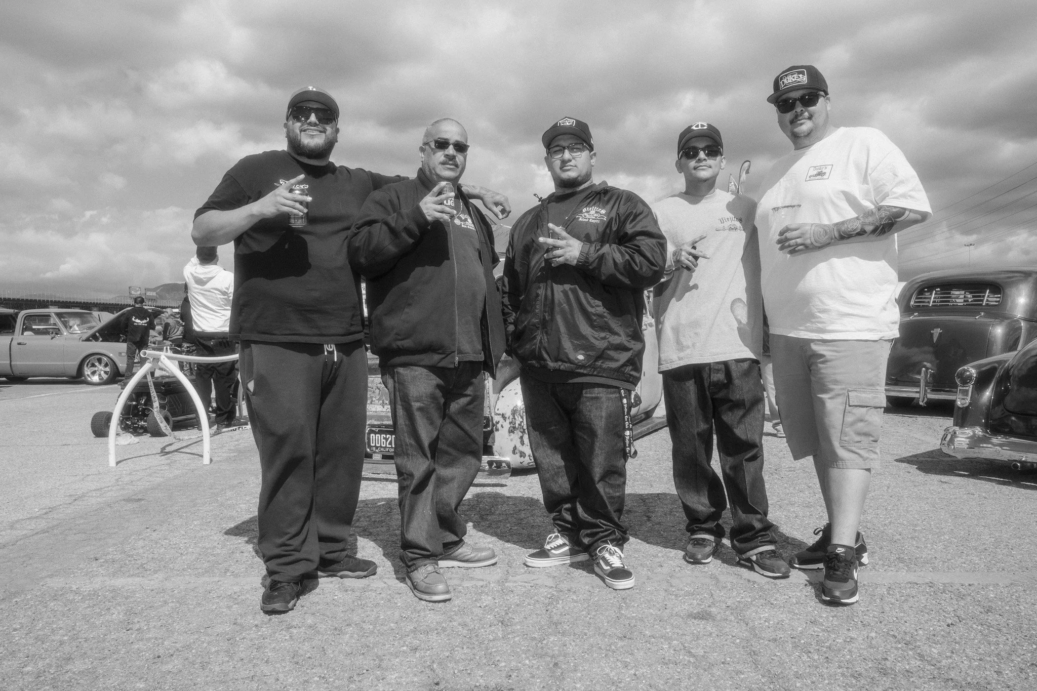 A group of six men standing outdoors in a lot with vintage cars in the background, some holding drinks and making hand gestures, under a partly cloudy sky.
