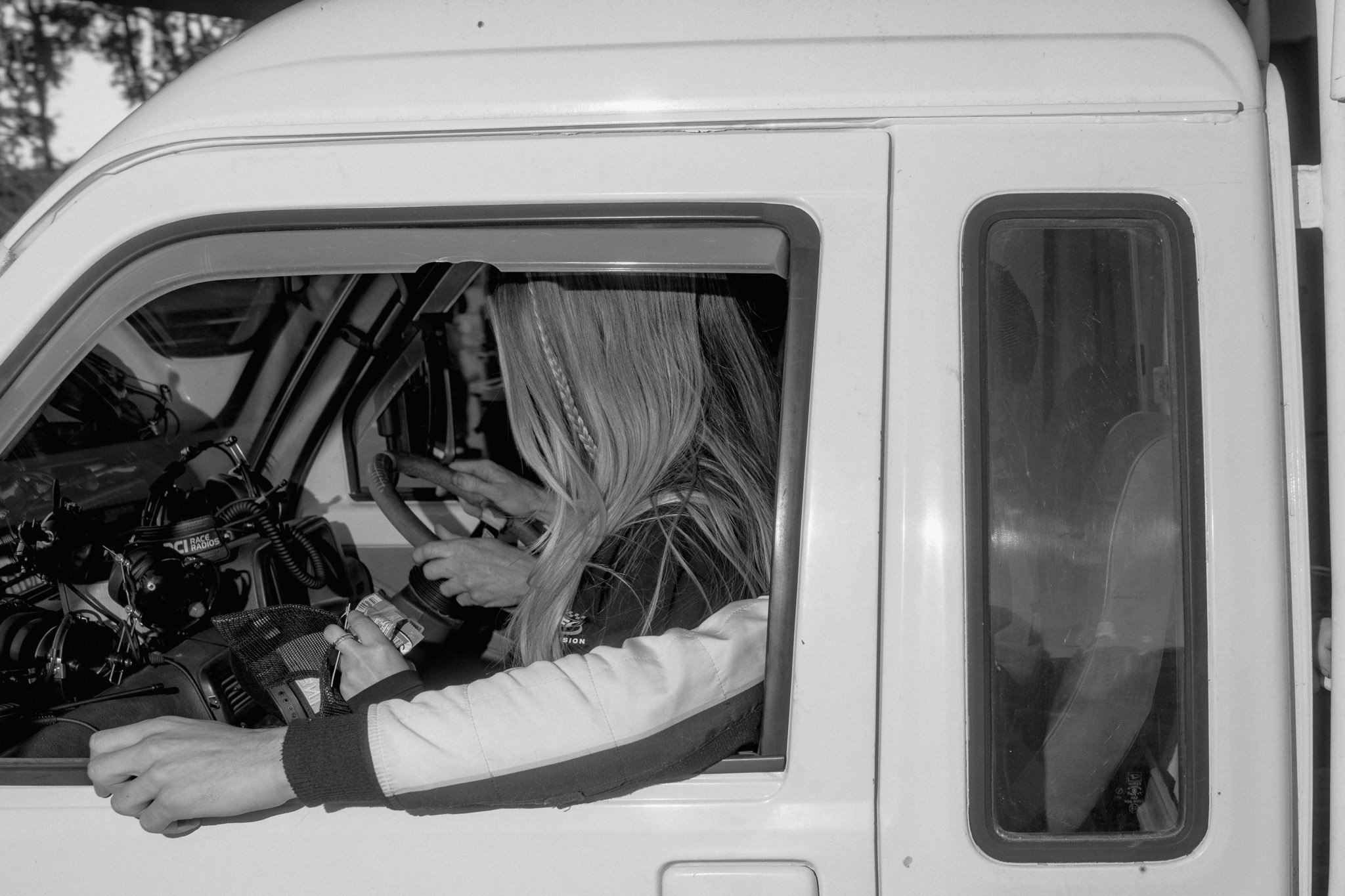 Black and white photograph of a young girl with long hair sitting on the lap of the driver, holding a smartphone, with the driver's left arm resting on the window ledge.