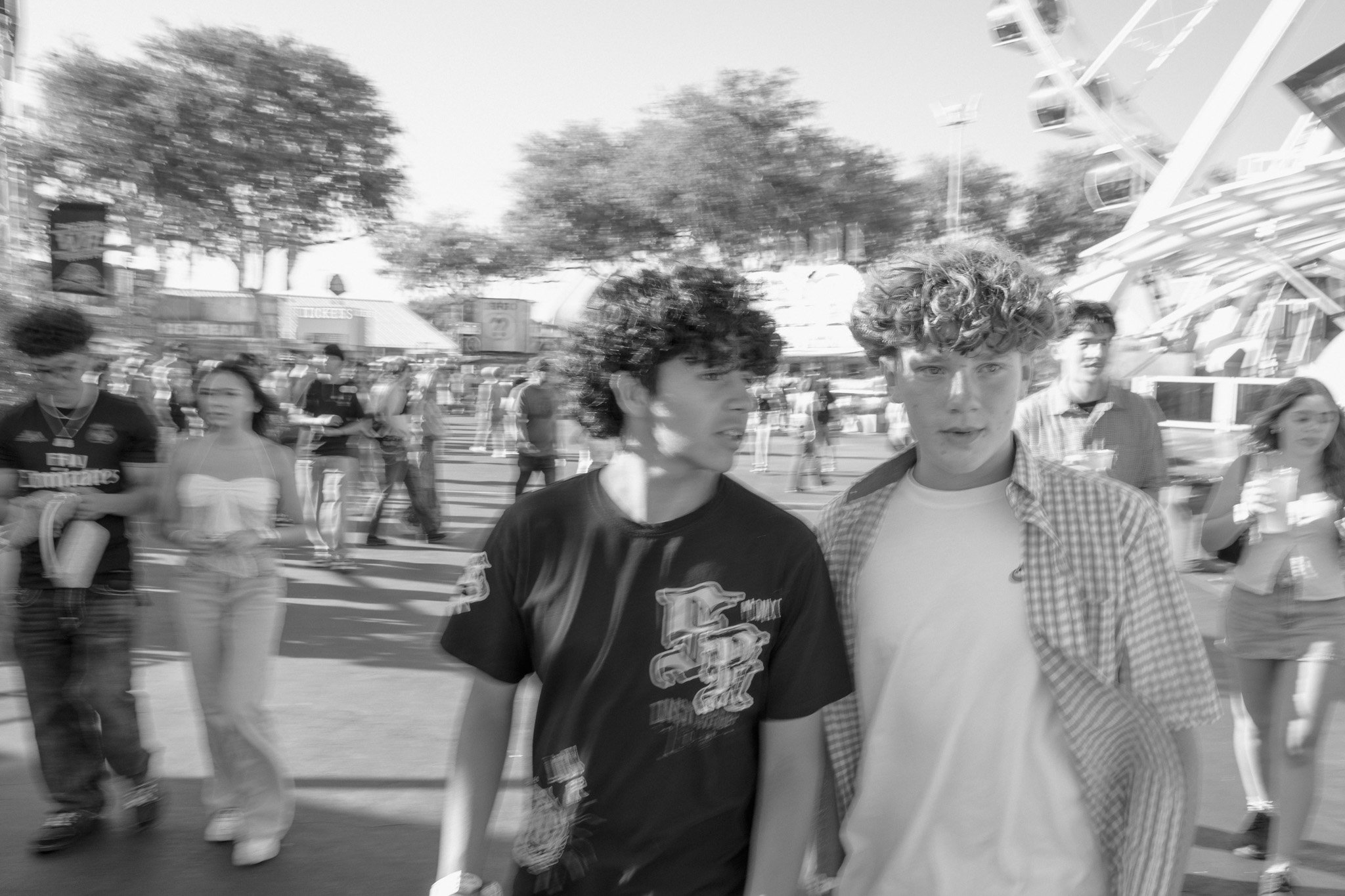 A black-and-white photo of two young men walking together at an outdoor event or fairground with a crowd, trees, and amusement rides in the background.