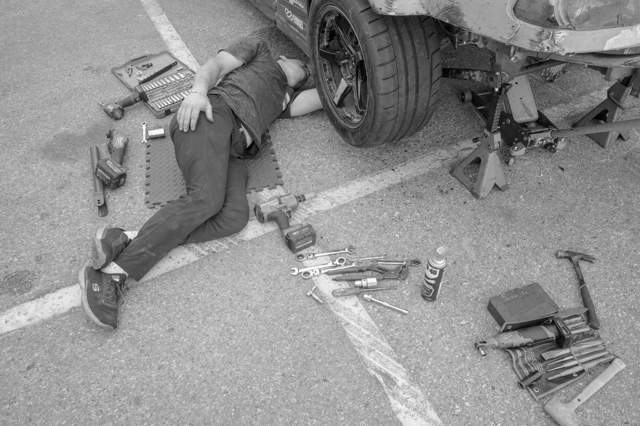 A man lying on the ground working under a car supported by jack stands in a parking lot, with various tools and equipment around him.