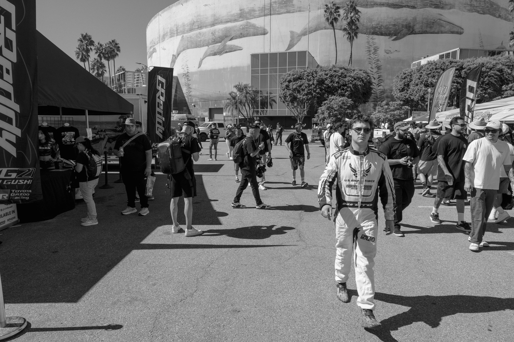A black and white photo of a crowd of people walking around an outdoor event, with several tents and flags, some people holding cameras and backpacks, and a large building with whale murals and palm trees in the background.