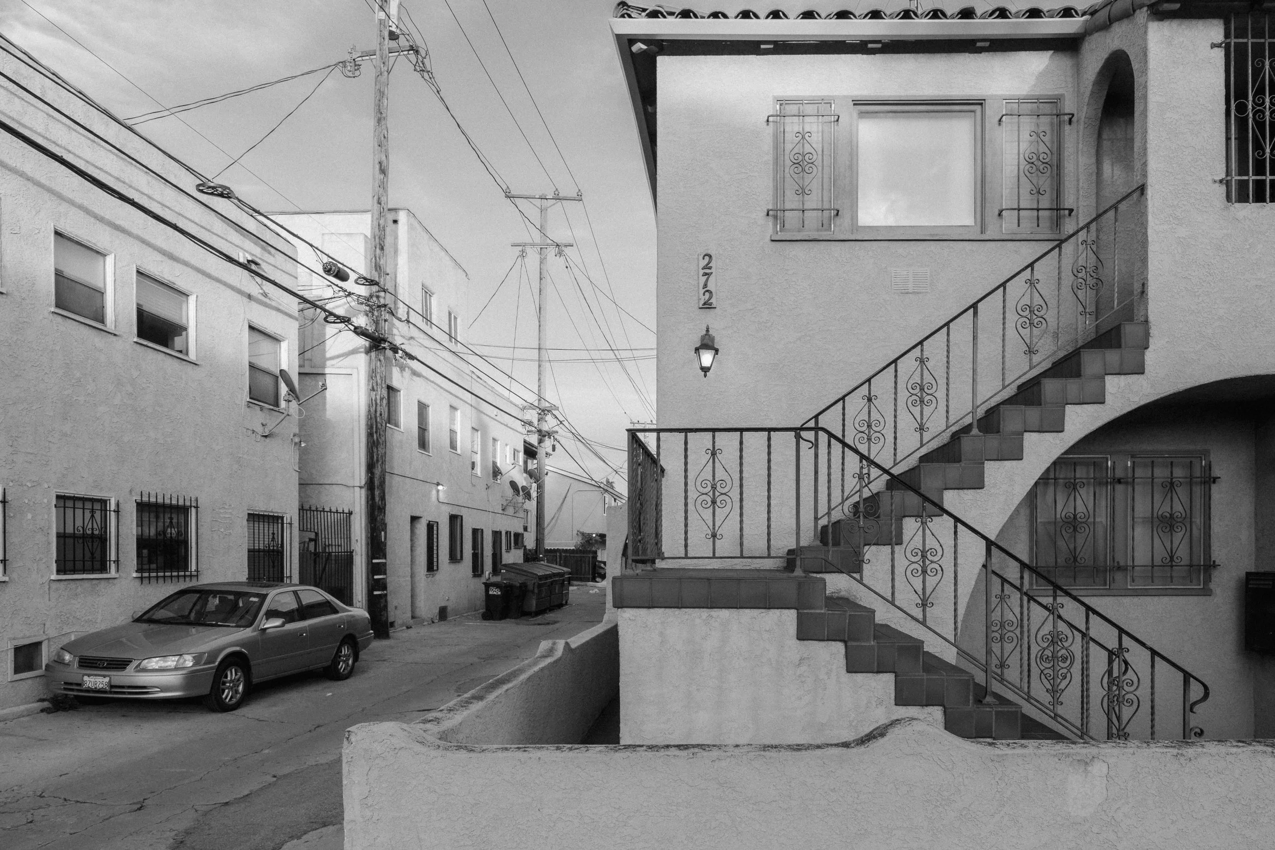 A black and white photograph of an urban residential alley showing a multi-storey building with stairs and railing, an parked car, utility poles and wires, and an overcast sky.