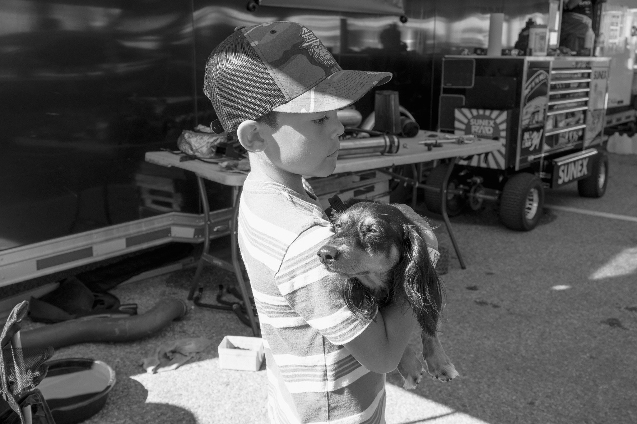 A young boy wearing a cap holding a small dog, possibly a Dachshund, at an outdoor event with tools and carts in the background.