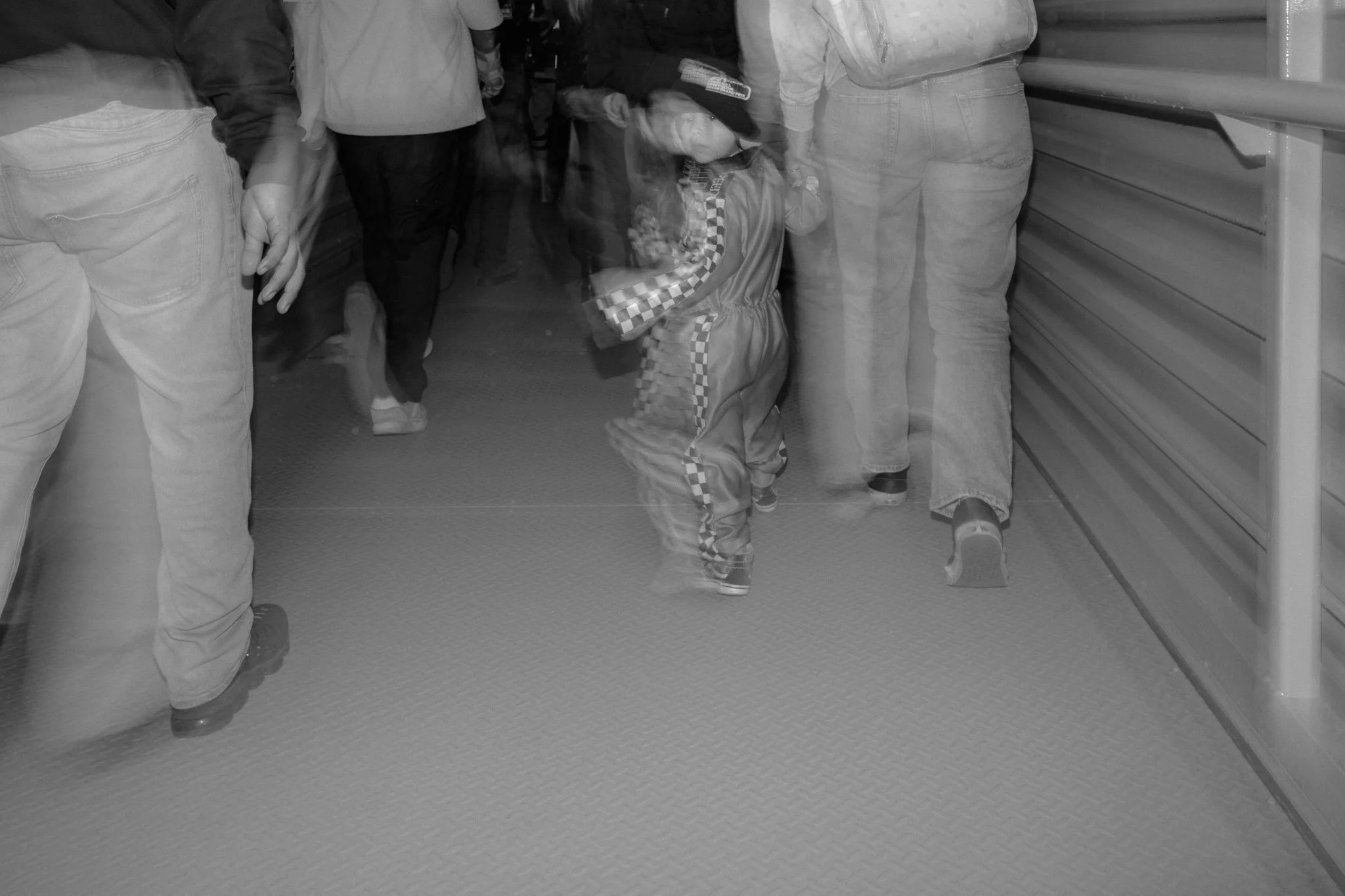A group of people walking through a crowded hallway, with a young child in a checkered racing jumpsuit costume and hat looking back at the camera.