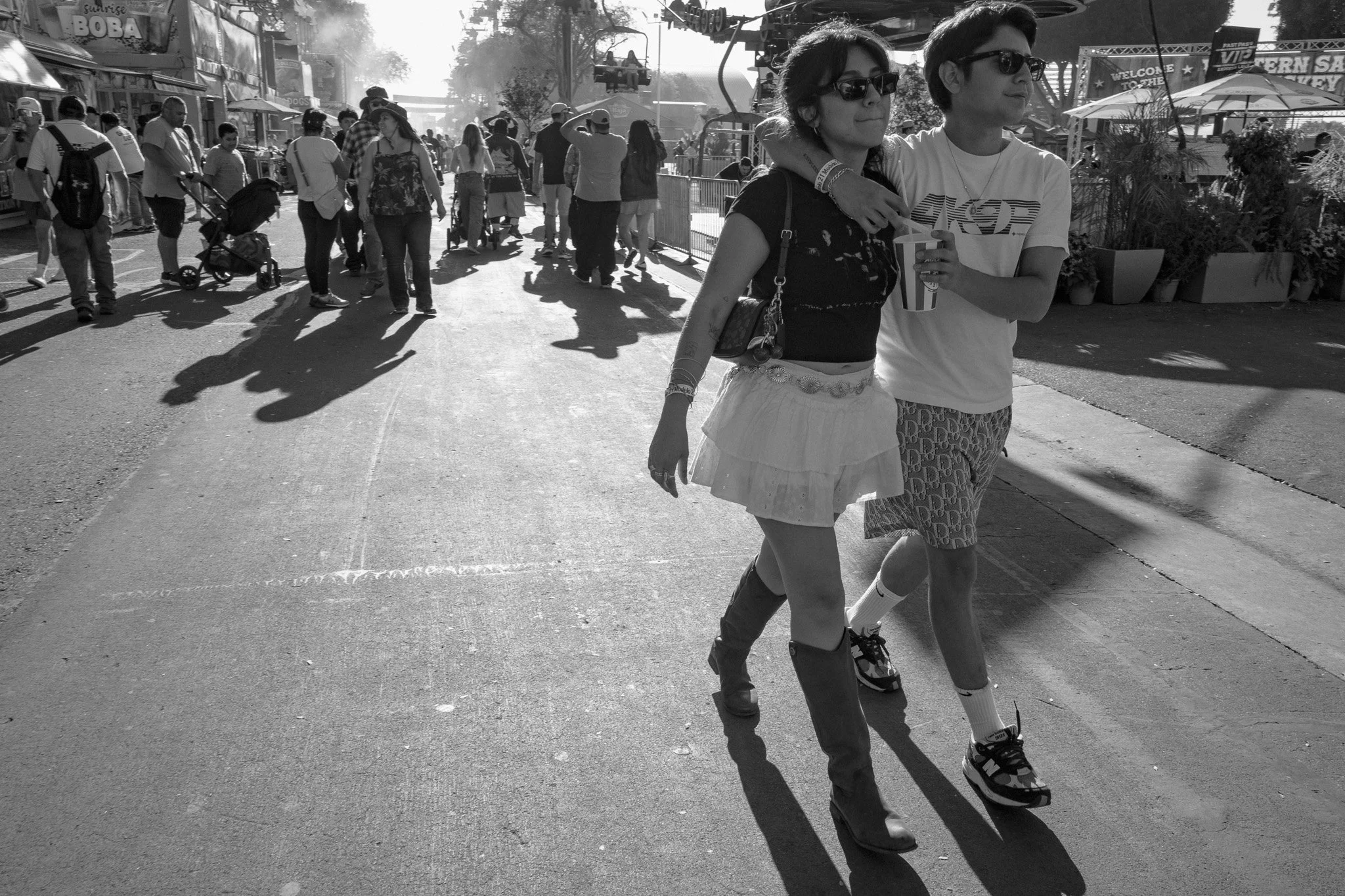 Two young women walk together at an amusement park, one with her arm around the other's shoulders, both wearing sunglasses, holding a drink, and smiling, with several people and amusement rides in the background on a bright day.