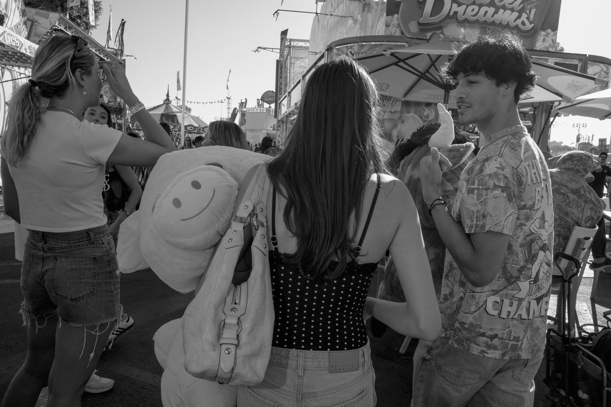Three people at a fair, two women and a man. The man is holding a food item, and the women are looking at him. The woman in the center has a large smiley face plush on her shoulder.