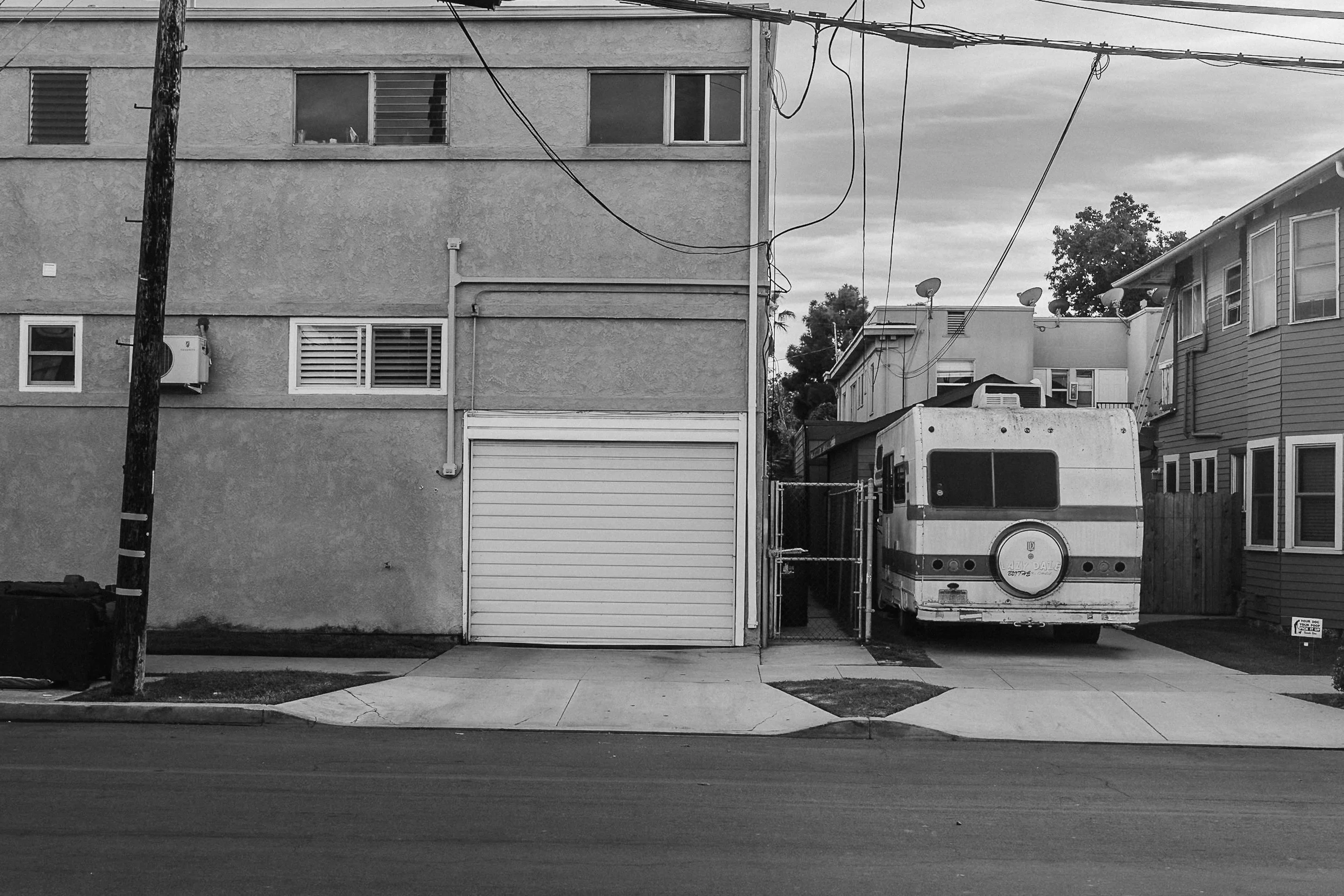 A black and white photo of a city street scene with a building on the left, showing windows, and a garage door. An RV is parked in a small driveway on the right, between two houses. Utility wires are overhead.