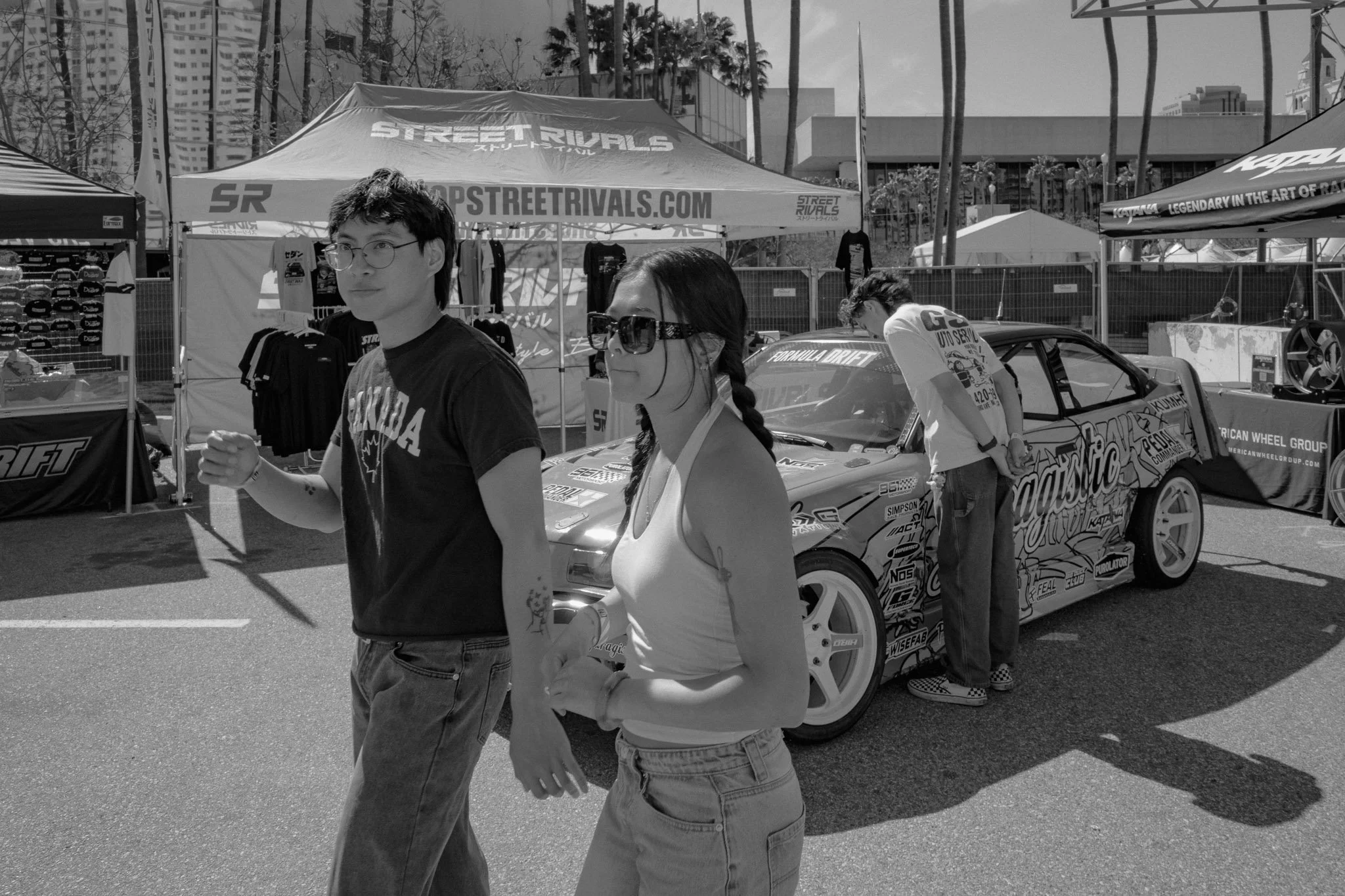 Two young adults, a man and a woman, walking past a race car at an outdoor automotive event. The man is wearing glasses and a black T-shirt, while the woman is wearing sunglasses, a tank top, and jeans. The car behind them is decorated with various s