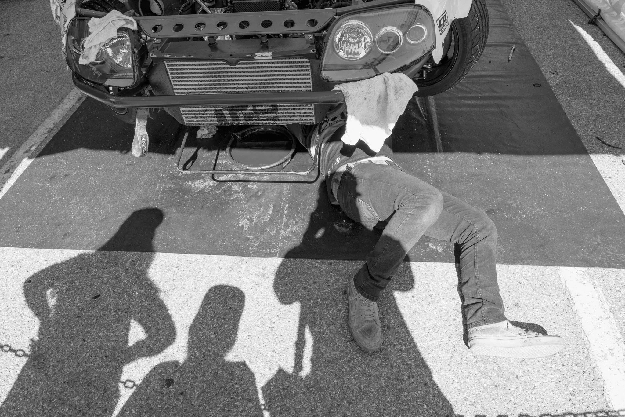 A person working under the front of a car on a parking lot, with tools and towels nearby, and shadows of other people cast on the ground.