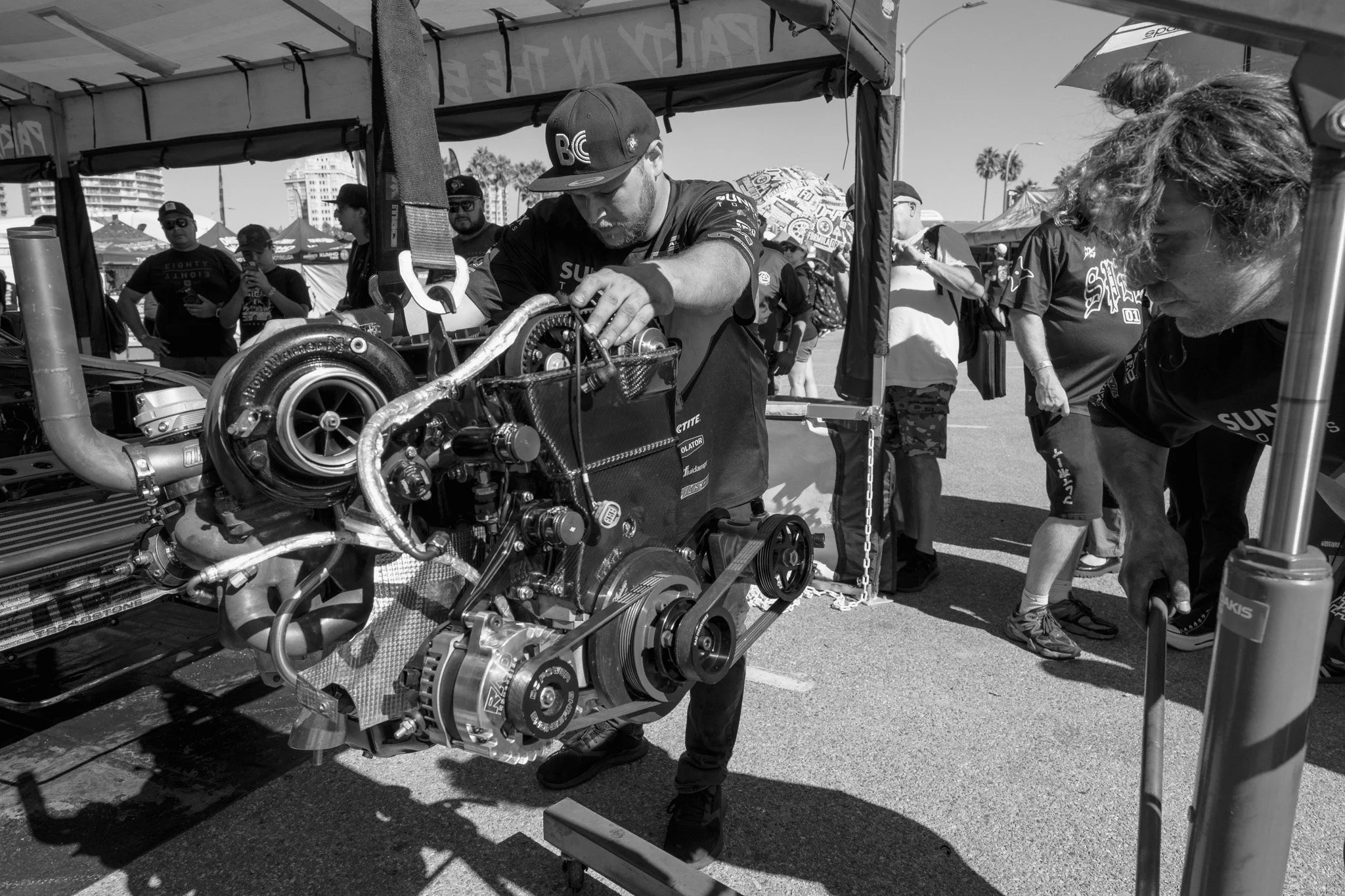 A black and white photo of a man working on a car engine at a racing event. Several people are observing in the background, some holding umbrellas and wearing event shirts, under tents in an outdoor setting.