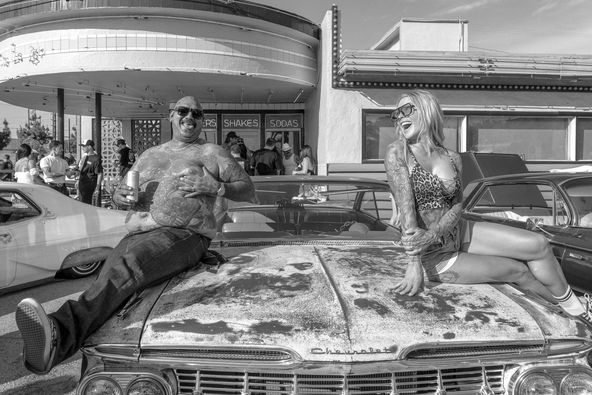 Two tattooed individuals, a man and a woman, sitting on the hood of an old Chevrolet car, smiling and laughing at a vintage car show outside a diner with a sign that reads 'Shakes, Sodas.' The man is shirtless, wearing sunglasses, and holding a can, 