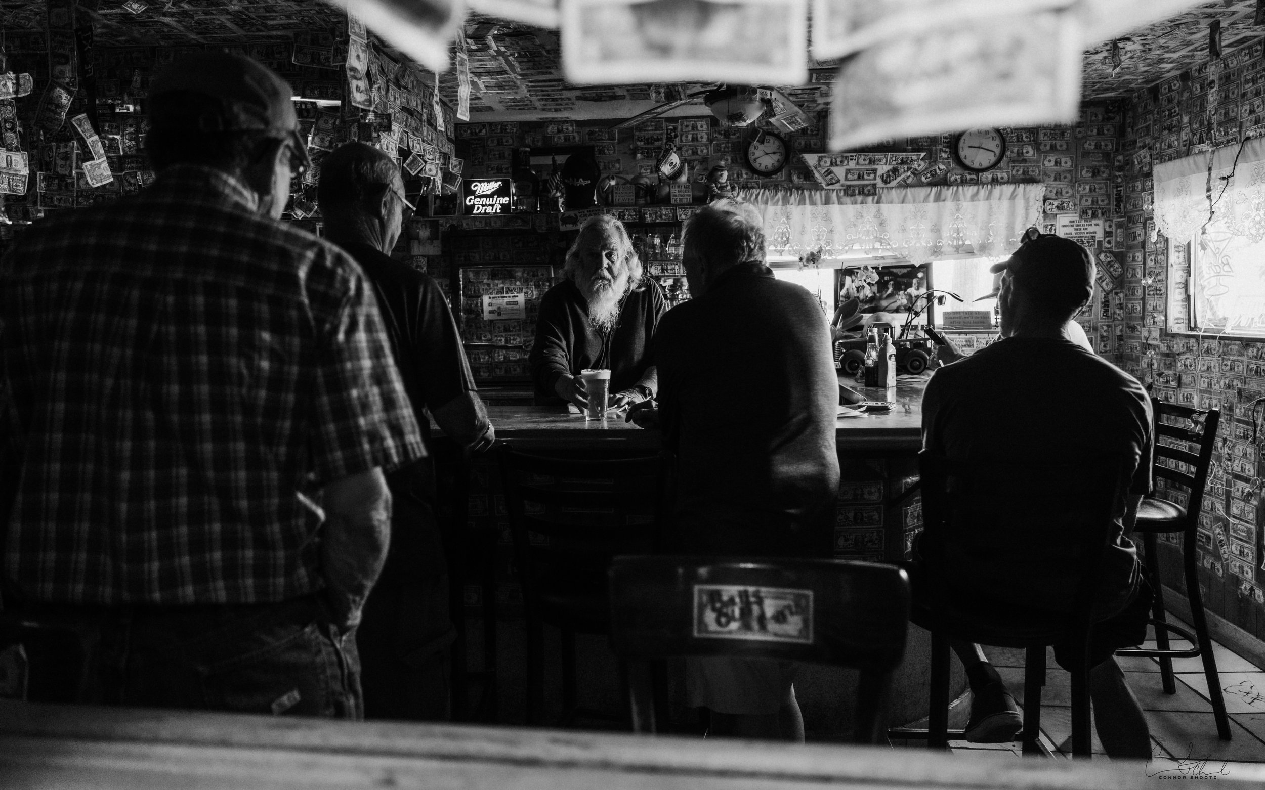 Black and white photo of five men gathered around a table in a bar or pub, with one man sitting at the center and four standing, engaged in conversation, with dollar bills and memorabilia on the walls.