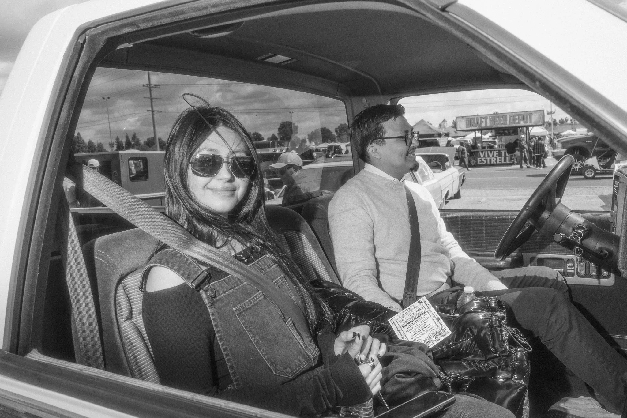 A woman and a man sitting inside a vehicle, with the woman in the passenger seat and the man driving, at an outdoor event with tents and people in the background.